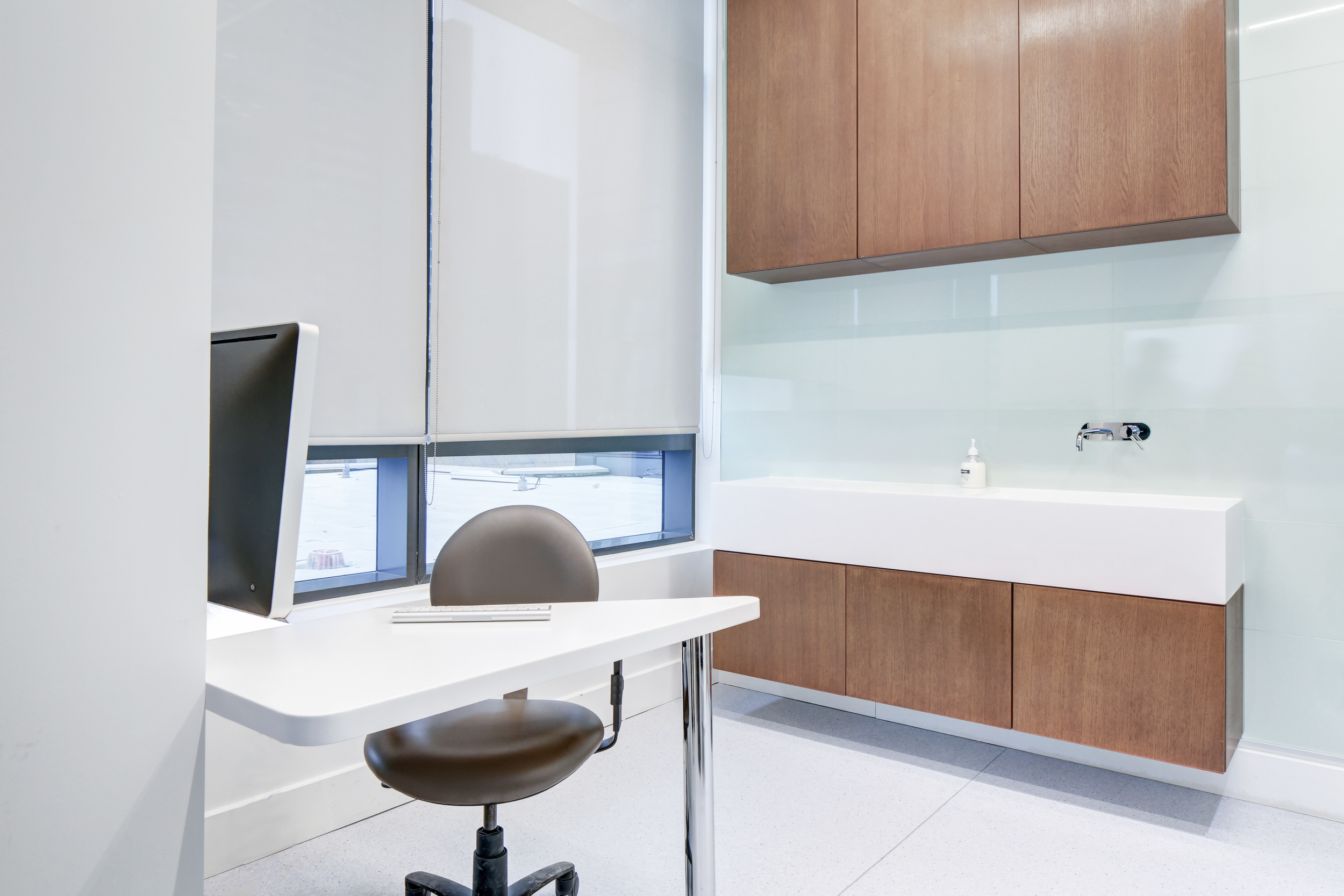 An office with a white desk, computer monitor, brown office chair, wooden cabinets, and a large window with blinds. healthcare, wellbeing. salutogenic design. wayfinding, human. inspiration for Toronto, Dallas, Texas, Miami, and Park City.