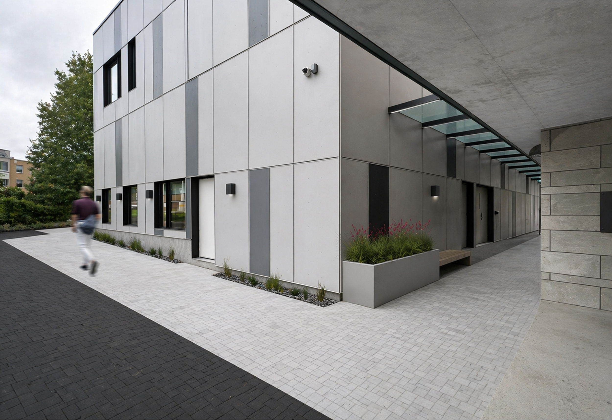 Modern building with gray exterior panels, large windows, security camera, and landscaped area with plants and flowers, sidewalk in front, overcast sky. Healthcare architecture. wellbeing and wayfinding. Inspiration for Toronto, Dallas and Park city.
