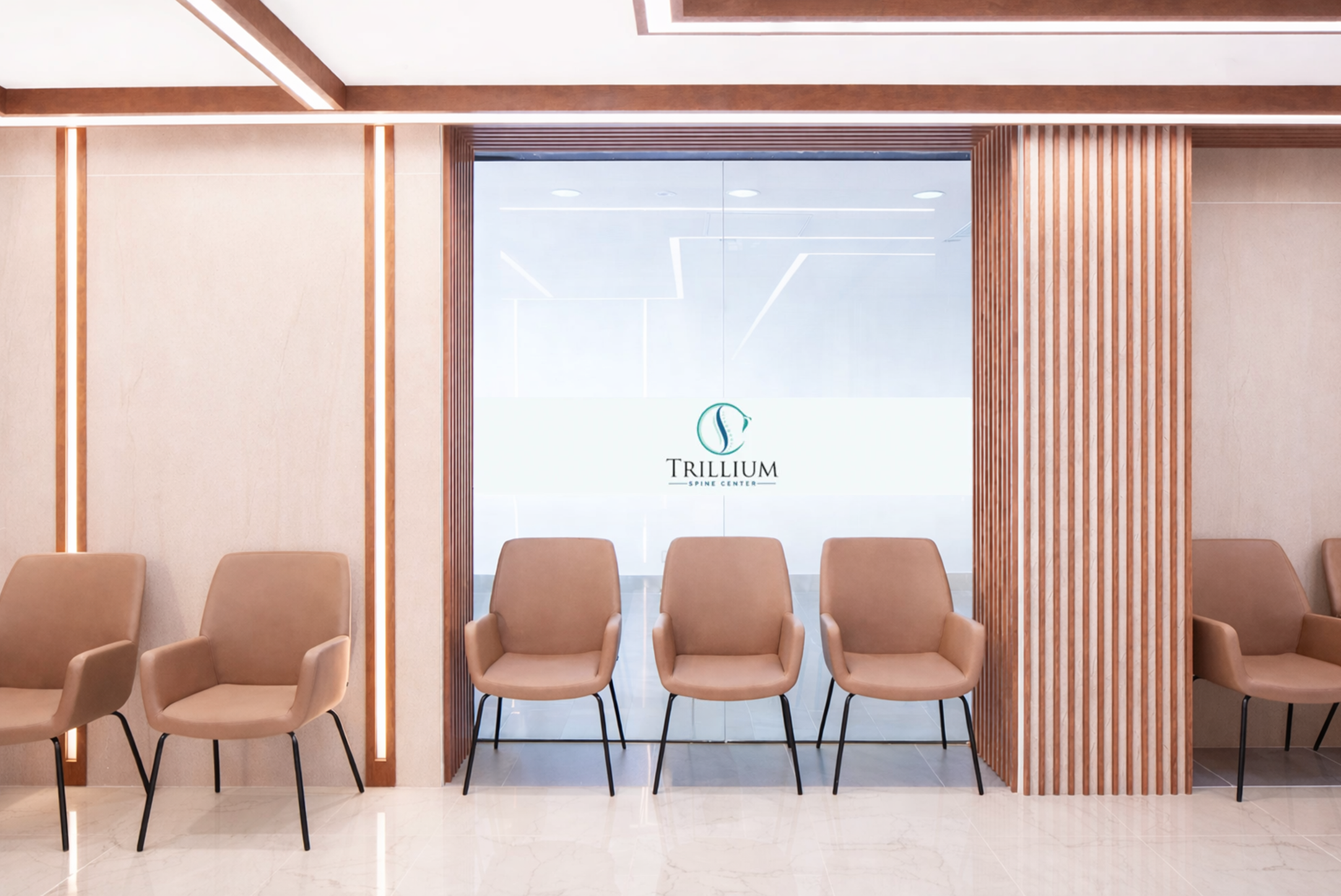 Waiting area with beige chairs in front of a glass wall that has the Trillium Spine Center logo, within a modern interior with wood accents and soft lighting. Healthcare. wellbeing and wayfinding. Inspiration for Toronto, Dallas, Park city.