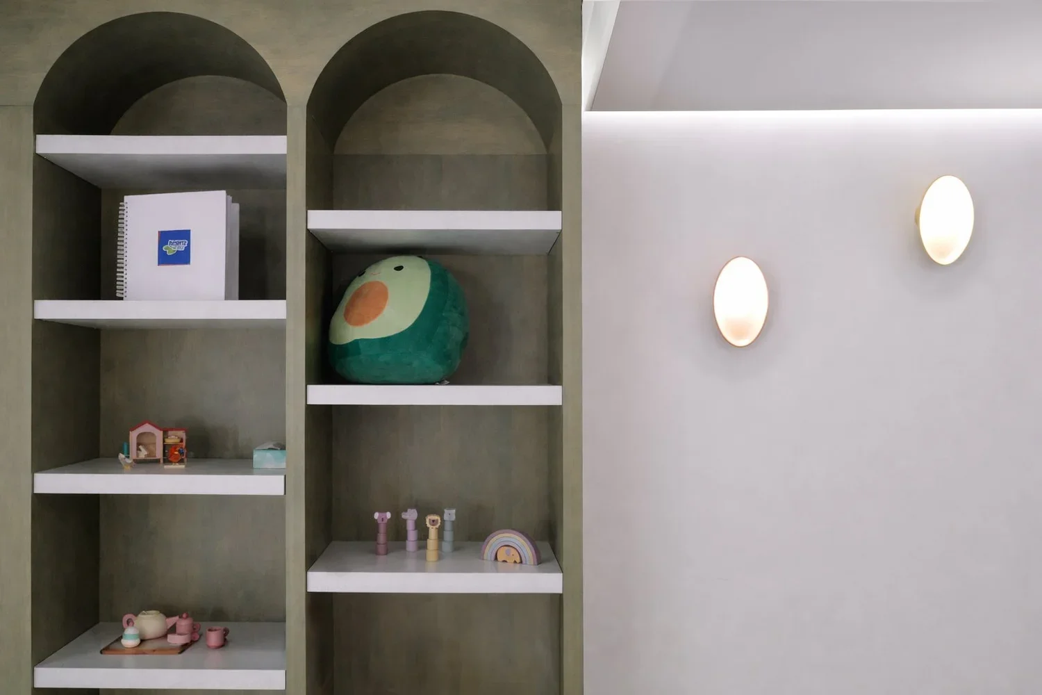 Interior view of a green bookshelf with white shelves containing toys, books, and decorations, next to a white wall with two oval wall lights.