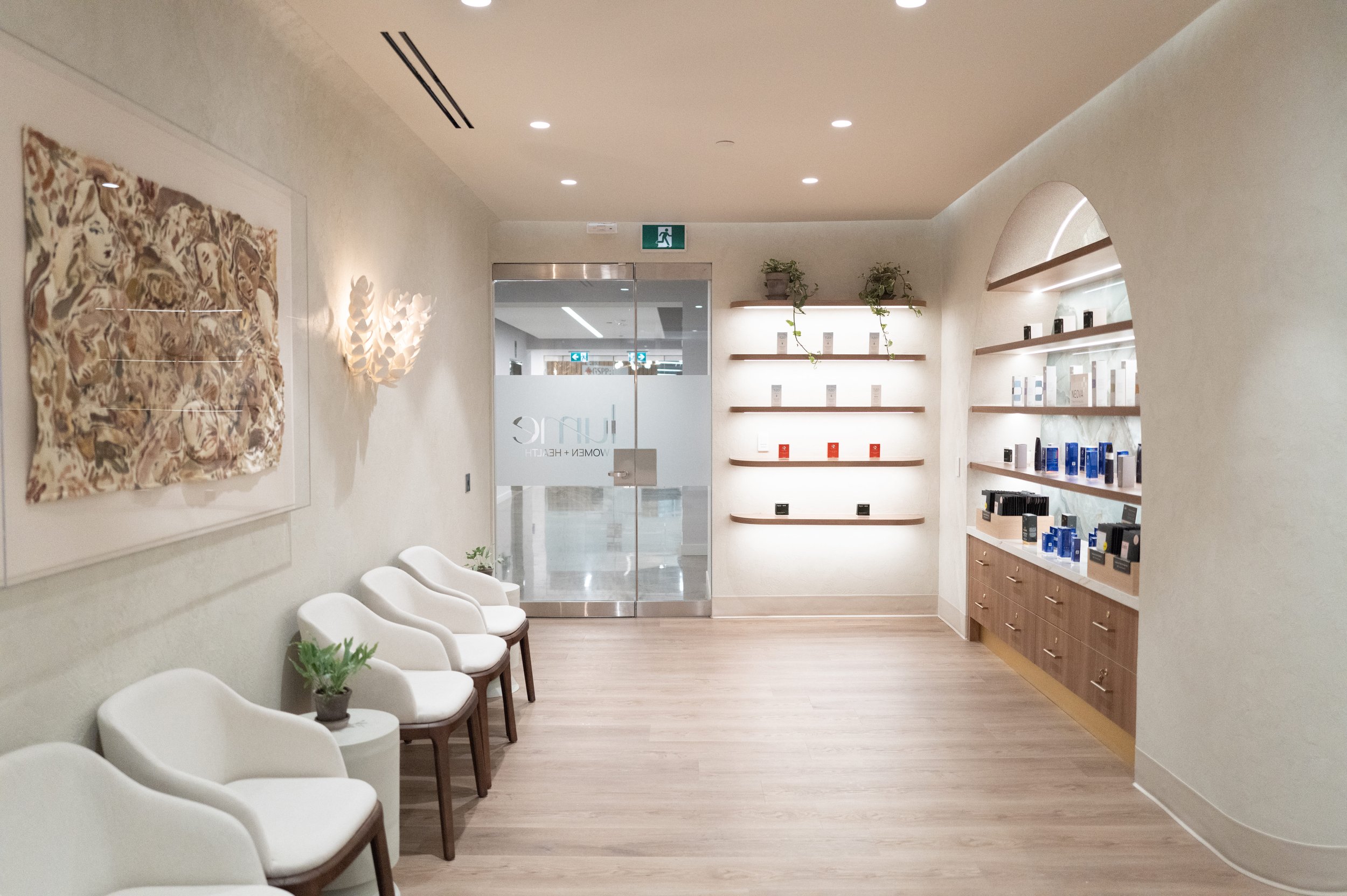 Minimalist waiting area with white chairs, a small table with a plant, wall art, and shelves with skincare products in a well-lit, modern interior