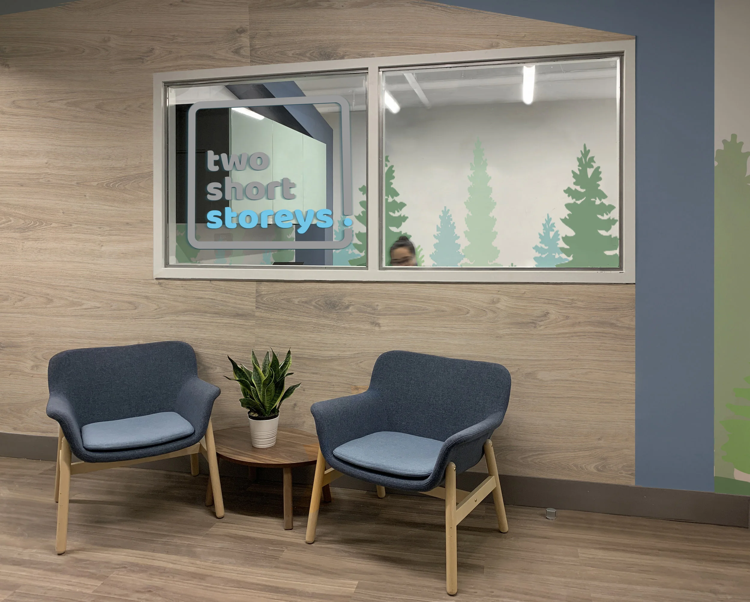 A waiting area with two blue chairs around a small round wooden table, a potted plant, and a window with a sign that reads 'two short storeys.' The window has a scenic forest mural with trees in the background.