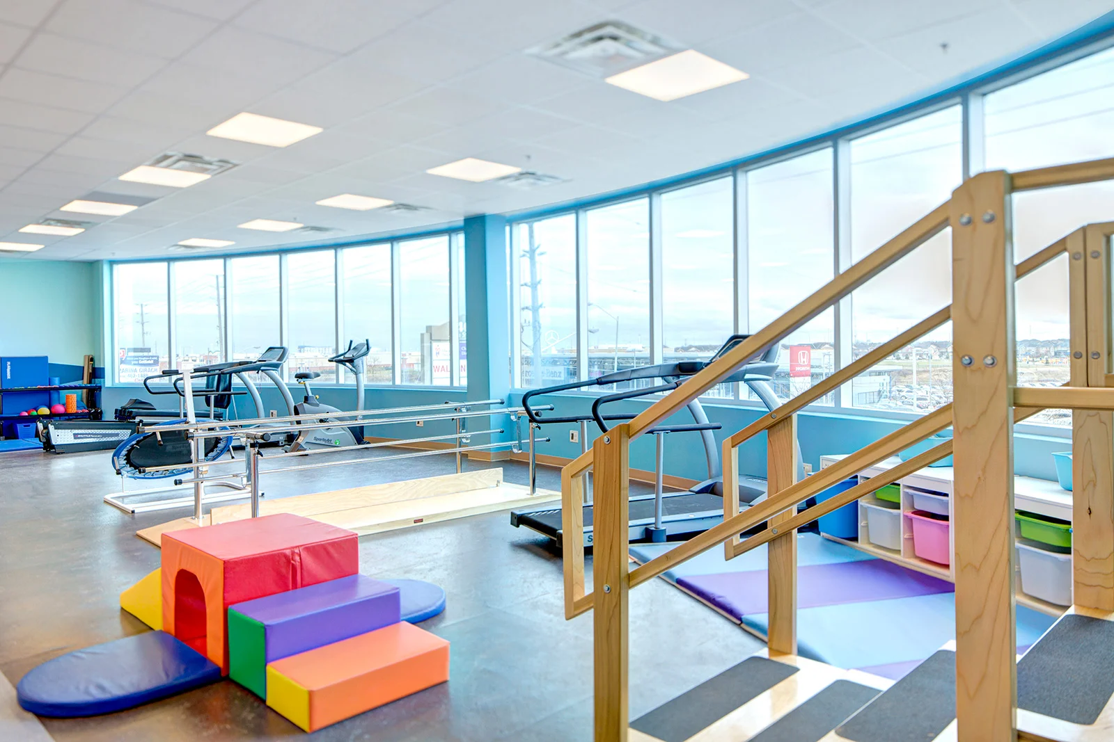 Bright, spacious physical therapy or gym room with large windows, exercise equipment including treadmills and elliptical, colorful foam blocks, padded mats, and a wooden staircase for exercises. healthcare.Toronto, Dallas, Texas, Miami and Park city.