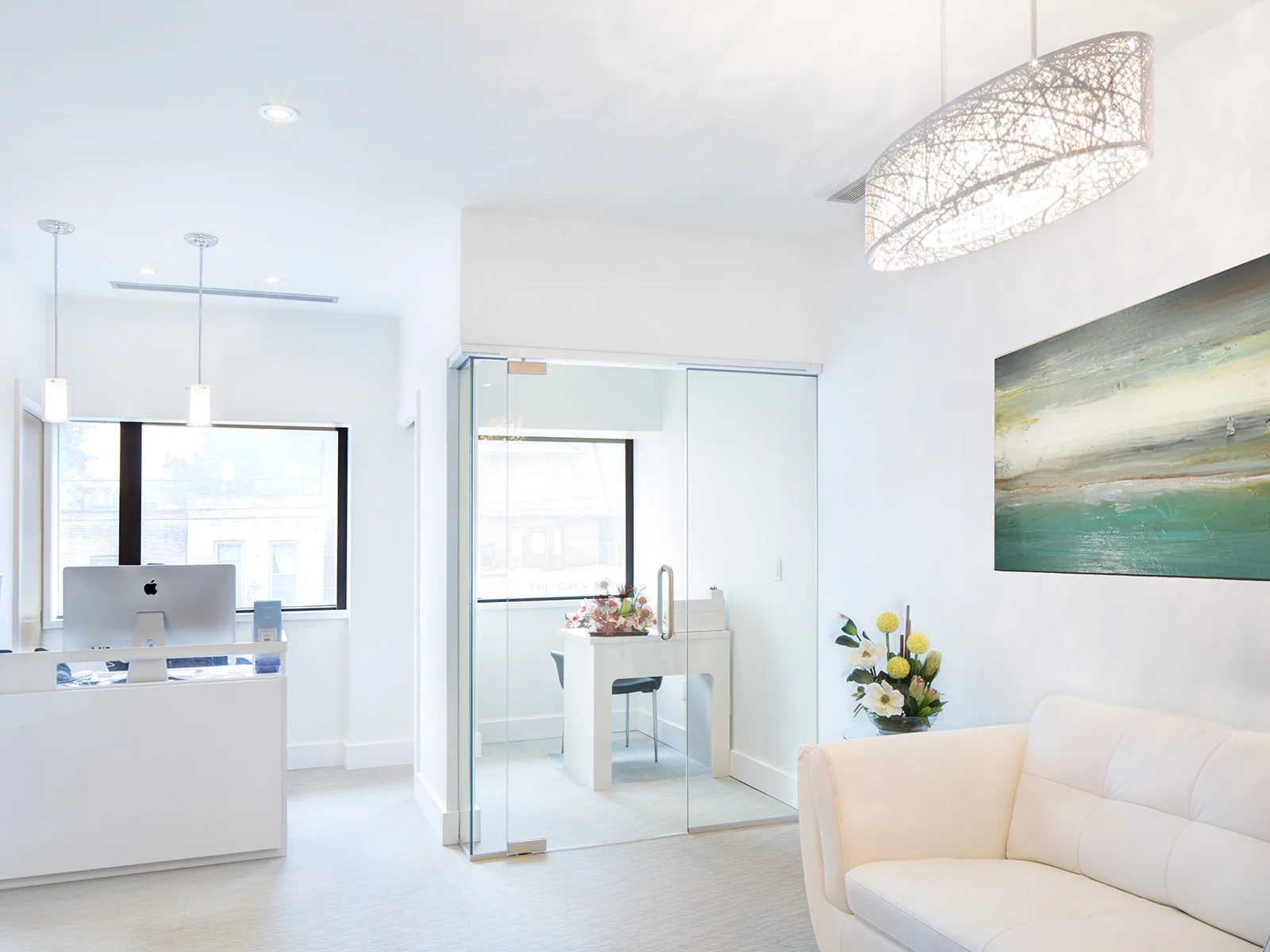 Bright, modern office space with white walls, a white desk, a sliding glass door, a white sofa with flowers, a large abstract painting, and decorative lighting fixtures. Healthcare. Inspiration for Toronto, Dallas, Texas, Miami and park city.