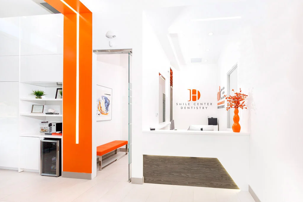 Modern dental office reception area with white walls, orange accents, a front desk with a computer, and a tall orange vase with orange flowers.