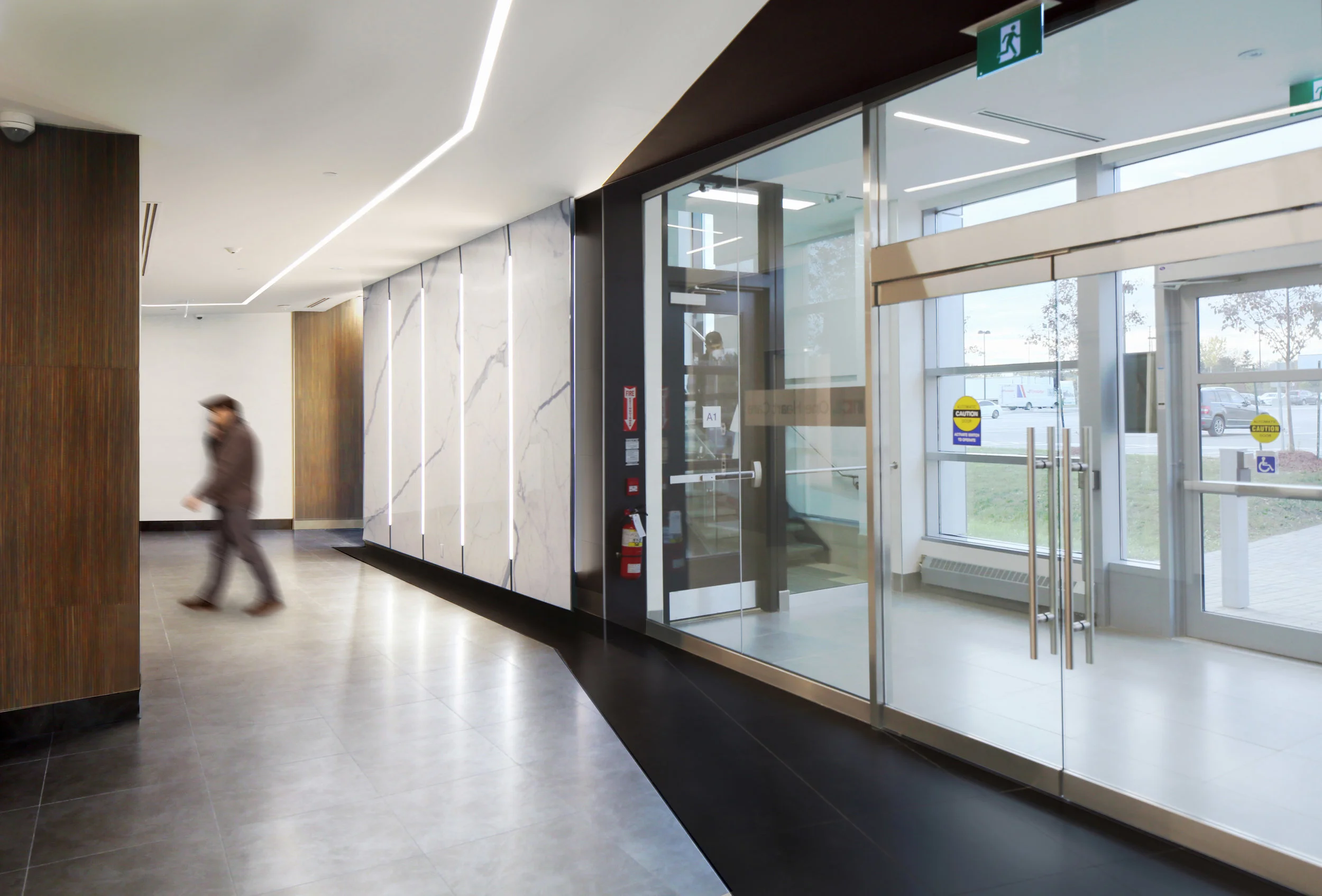 Modern building interior with a man walking and glass doors leading outside, emergency exit sign above the doors. Wellbeing and flow, salutogenic design. Healthcare architecture. Good inspiration for Toronto, Dallas and Park City.