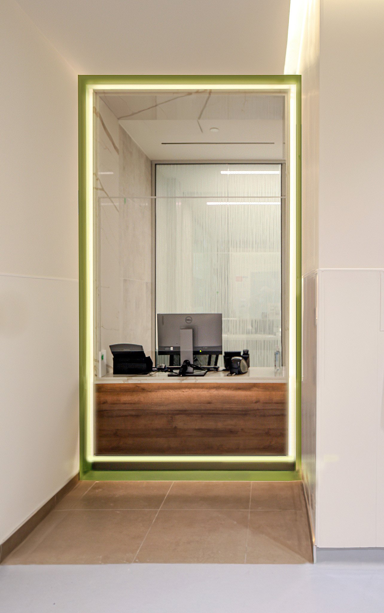 A mirror with green trim reflecting a workspace with a computer monitor, keyboard, and various office items, set against a glass wall with vertical blinds and a ceiling panel. Healthcare architecture inspiration for Toronto, Dallas and Park City.