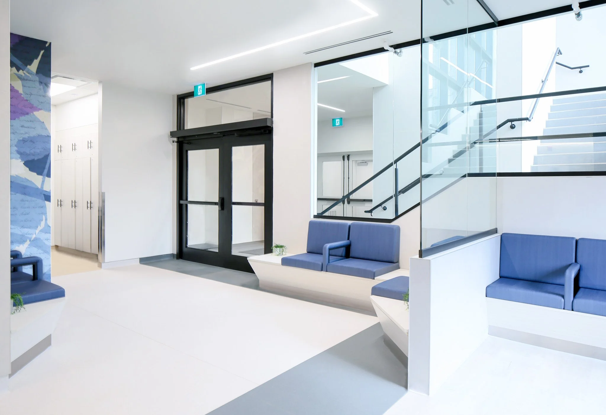 Modern waiting area promotes wellness and healthcare with blue cushioned benches and glass staircase in a bright, minimalistic building lobby. Designed to have good flow, promotes wayfinding. Good inspiration for Toronto, Dallas and Park City.