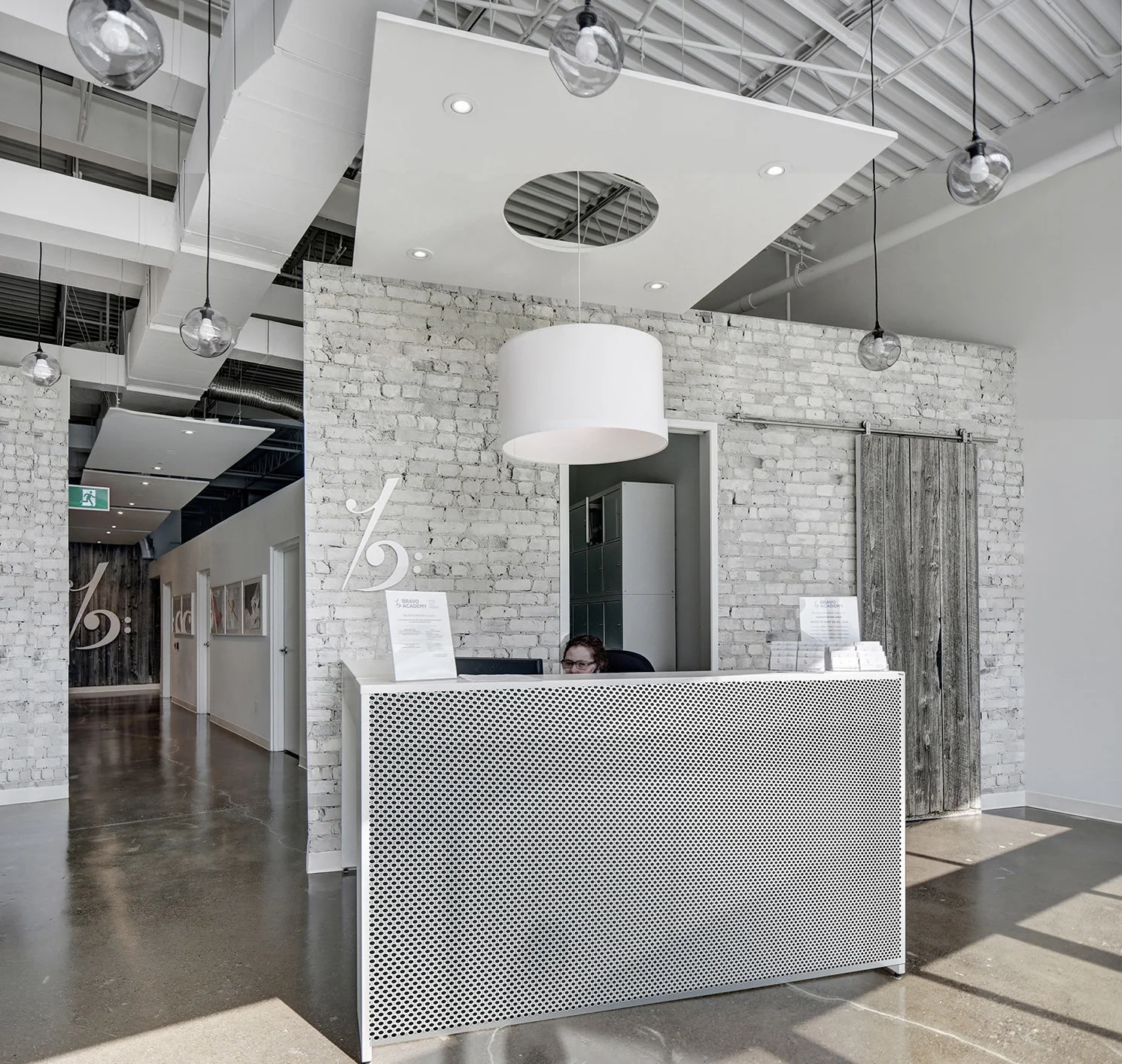 Modern reception area with white brick wall, a woman sitting behind a white perforated desk, and a large white cylindrical light fixture hanging from the ceiling. Glass pendant lights are suspended from the ceiling, and there's a sliding barn door ma