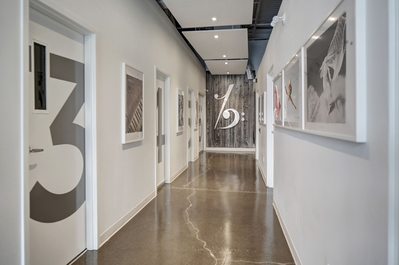 A modern hallway with white walls, framed artwork, polished concrete floors, and a wood accent wall at the end with a large white percent sign. Education architecture. wellbeing and flow. Inspiration for Toronto, Dallas, Texas, Miami, and 