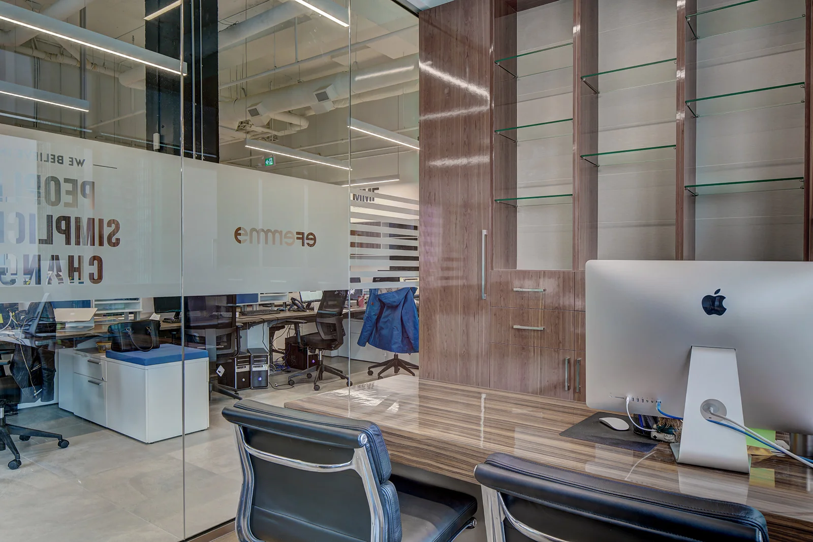 An office seen through a glass wall, with a wooden cabinet, a desk with an iMac, chairs, and an office space with computers and desks visible in the background. Education Architecture.  wayfinding.  Toronto, Dallas, Park city, Texas, Miami.