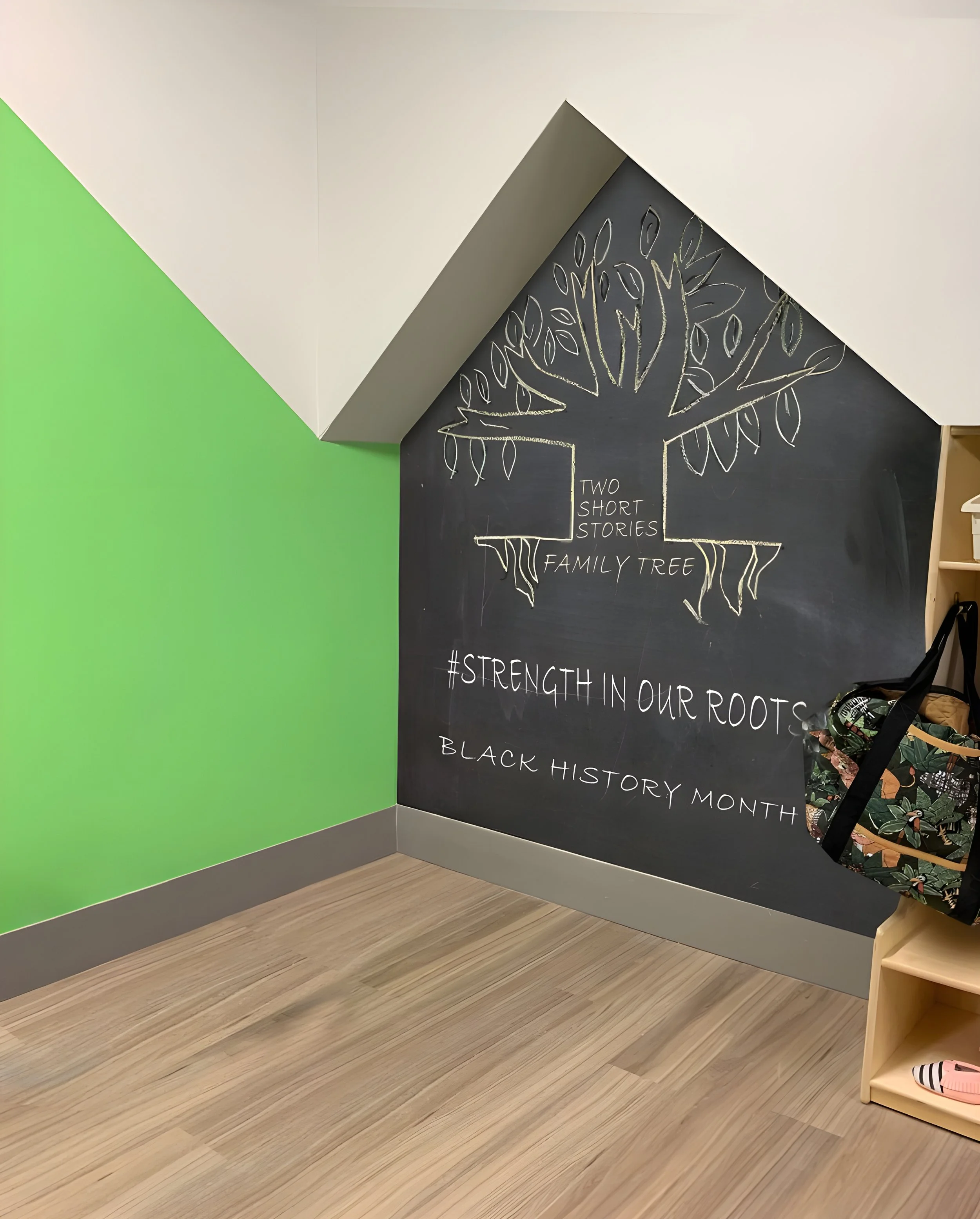 Corner of a room with green and black walls, a chalkboard with a chalk drawing of a tree, and a small wooden shelf with a plant and a pink sandal.
