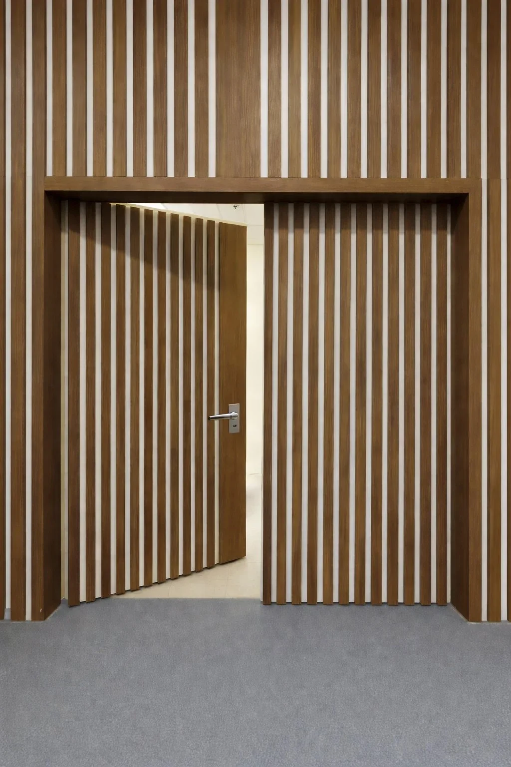 Wooden slat wall with an open door leading to a hallway with beige walls and a grey carpeted floor.