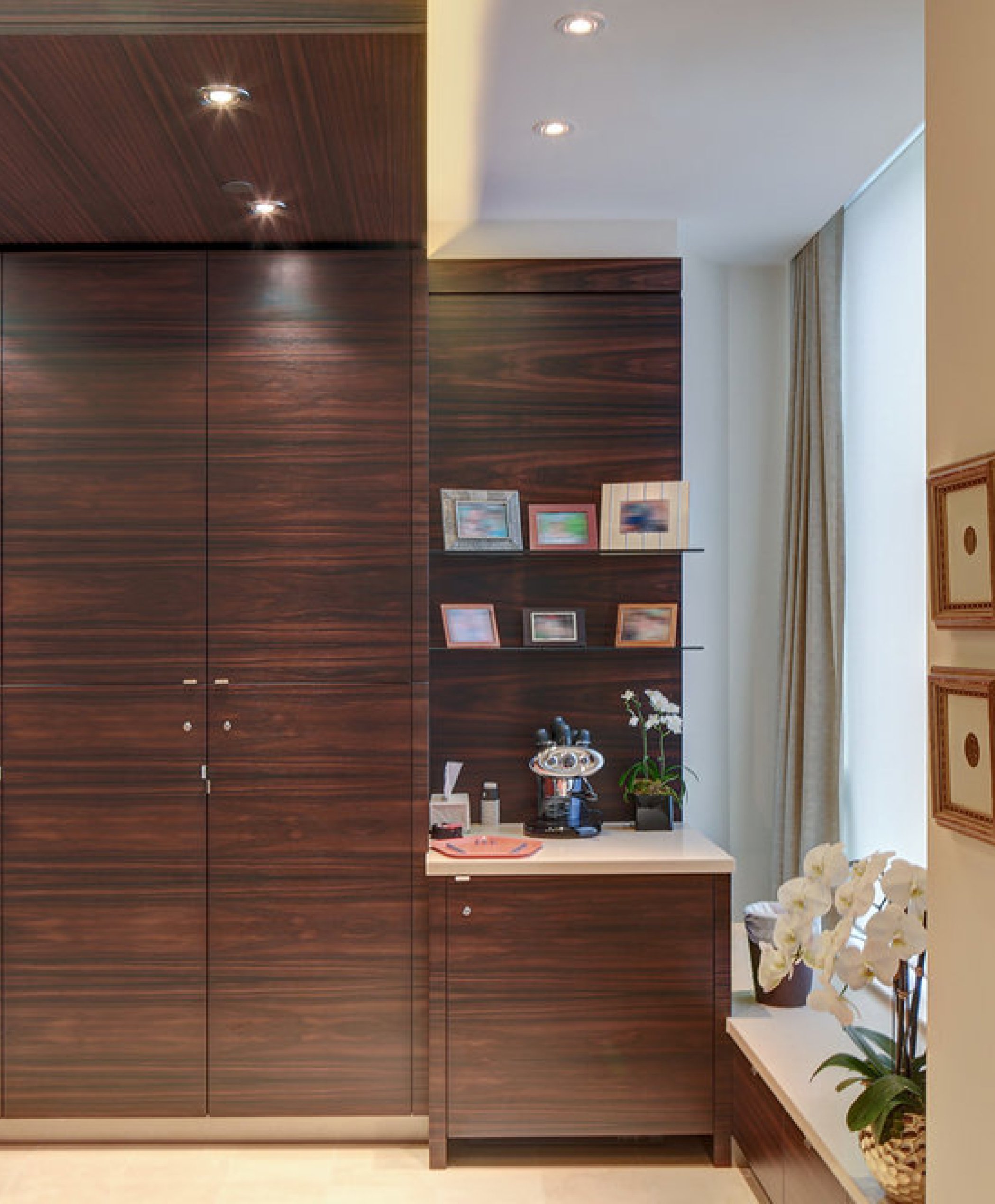 Interior view of a room with a large wooden cabinet, a small countertop with framed photographs, a flower plant, and a window with curtains, in a well-lit space.