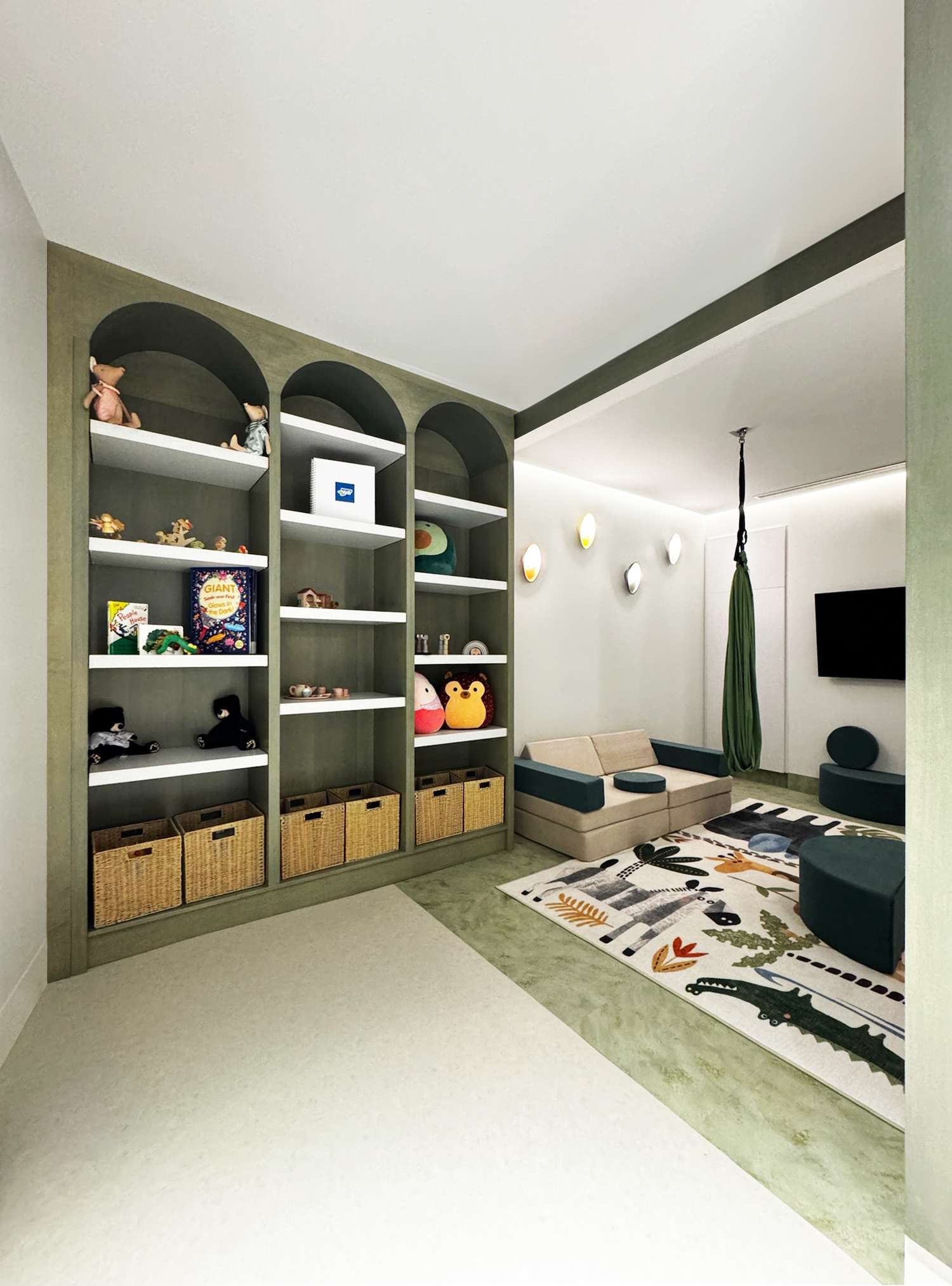 Interior of a children's playroom with a green and white color scheme, a bookshelf filled with toys and books, a beige sofa, a colorful area rug with animal and nature designs, a wall-mounted television, and a hanging fabric swing.