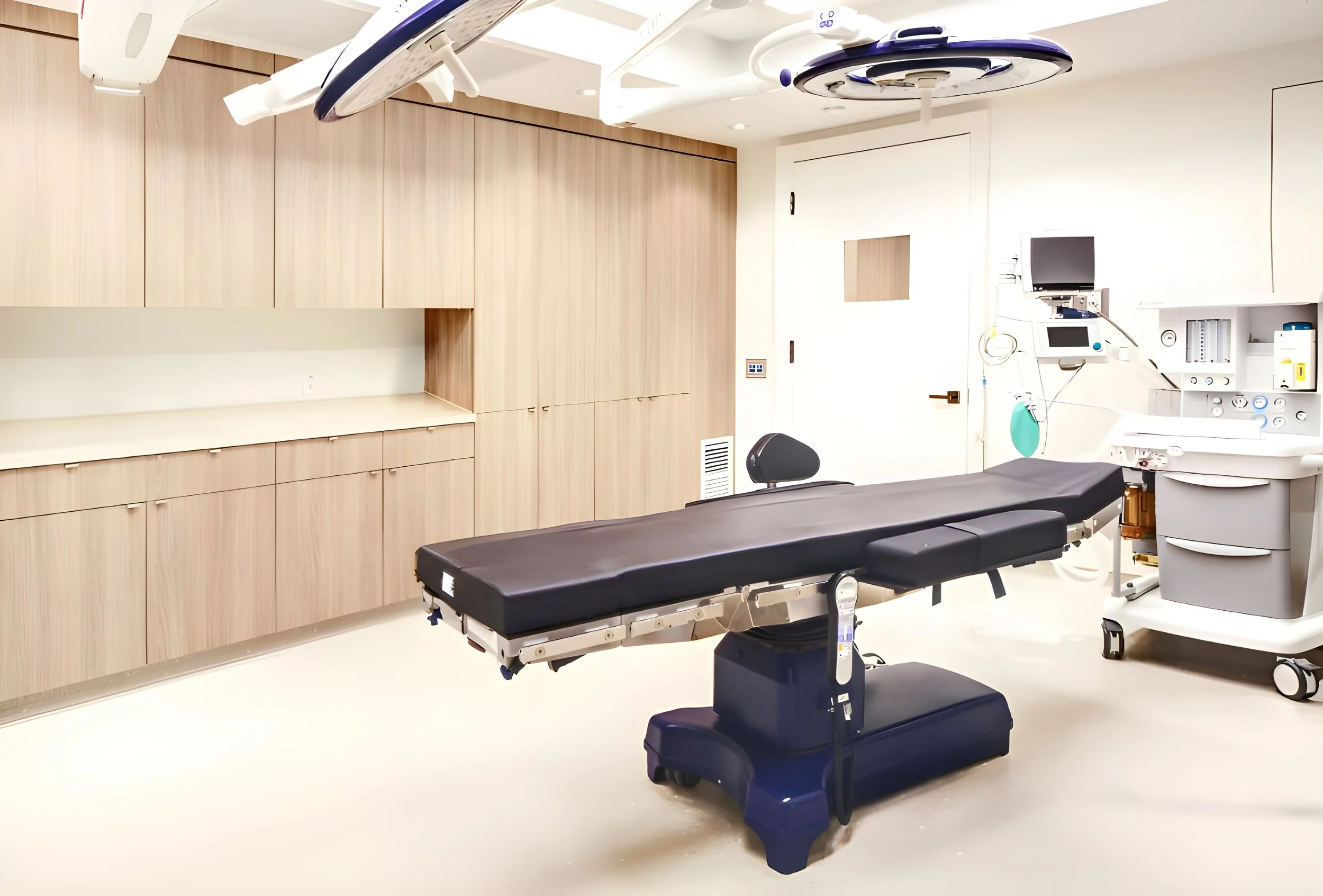 A hospital operating room with a medical examination table, surgical lights, cabinets, and medical equipment.