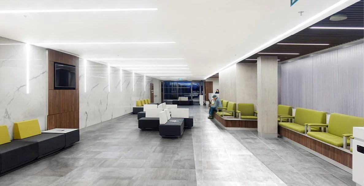 Modern waiting area with green and white chairs, gray floors, marble walls, and a television mounted on the wall. A person is sitting in one of the chairs near the window. Wellbeing and flow. Good inspiration for Toronto, Dallas and Park City.