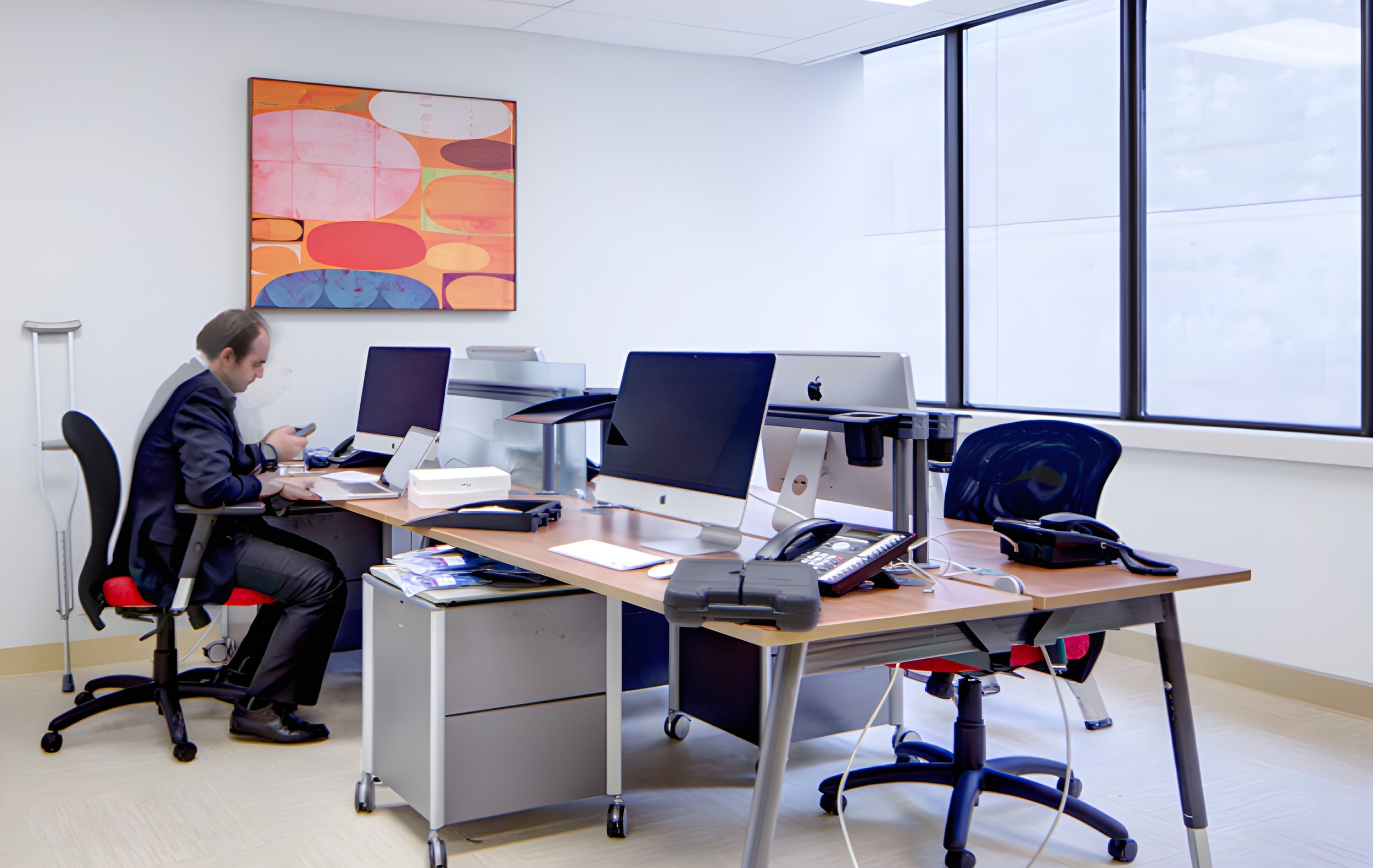 An office with a man sitting at a desk, working on a phone. There are multiple computer monitors, office supplies, and papers on the desk. Large windows are on the right side, and abstract colorful artwork hangs on the white wall.