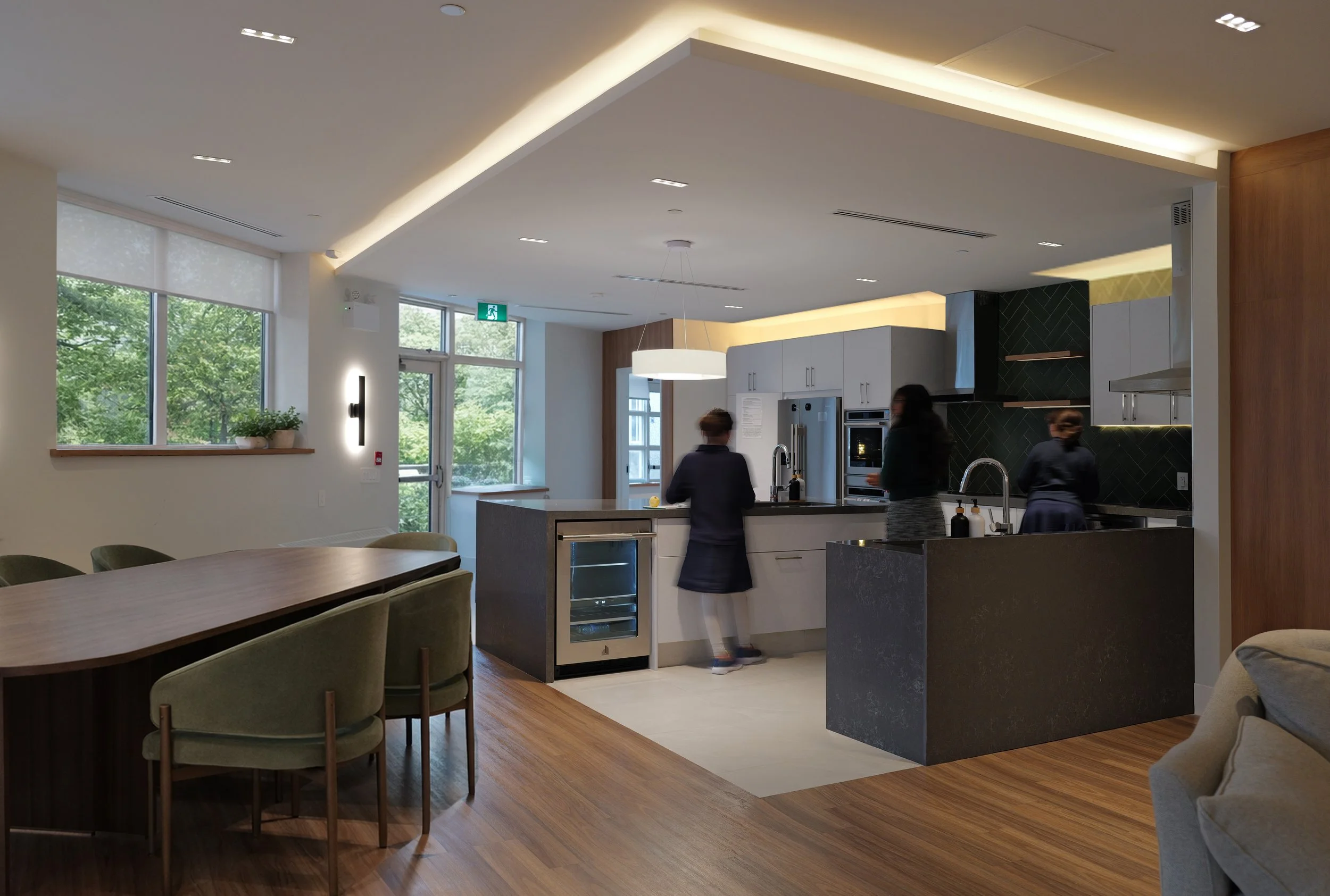 Modern, brightly lit kitchen with people preparing food, large windows with greenery outside, and wooden floors.