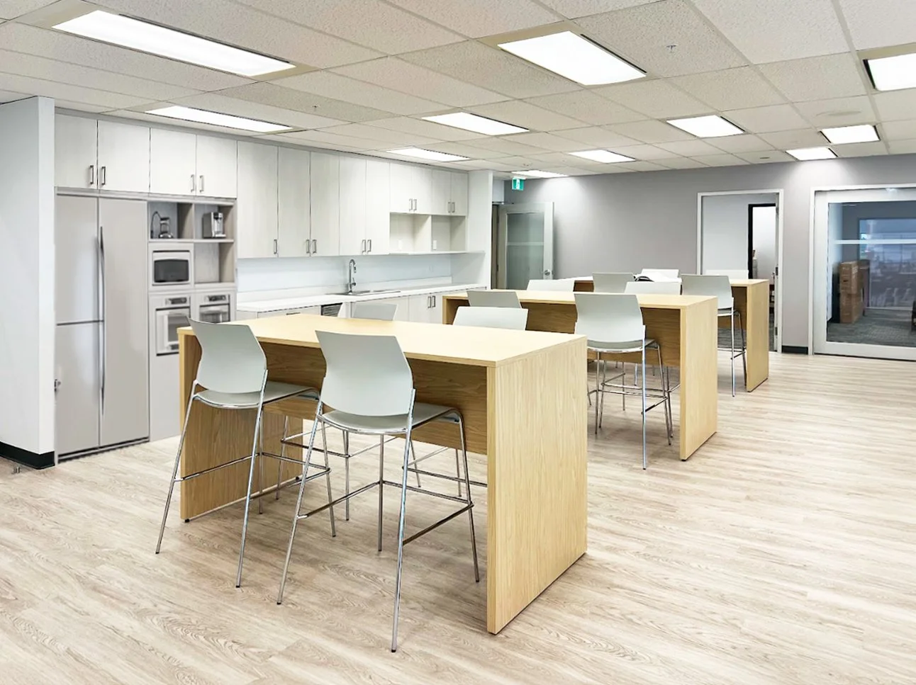 A modern, empty kitchen and break area with wooden tables, white chairs, white cabinetry, and kitchen appliances like a refrigerator and microwave, illuminated by ceiling lights.