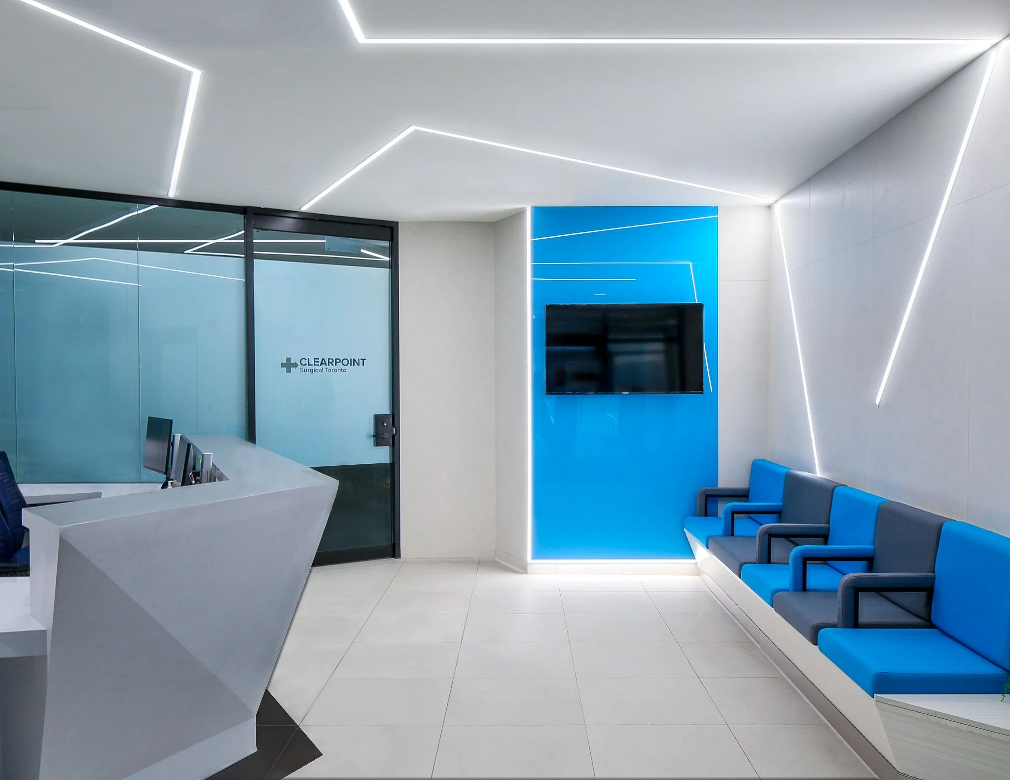 Modern medical reception area with white walls, blue accents, row of blue chairs, a white reception desk, and a wall-mounted television screen. Salutogenic design, promotes health and beauty. Good inspiration for Toronto, Dallas and Park City.