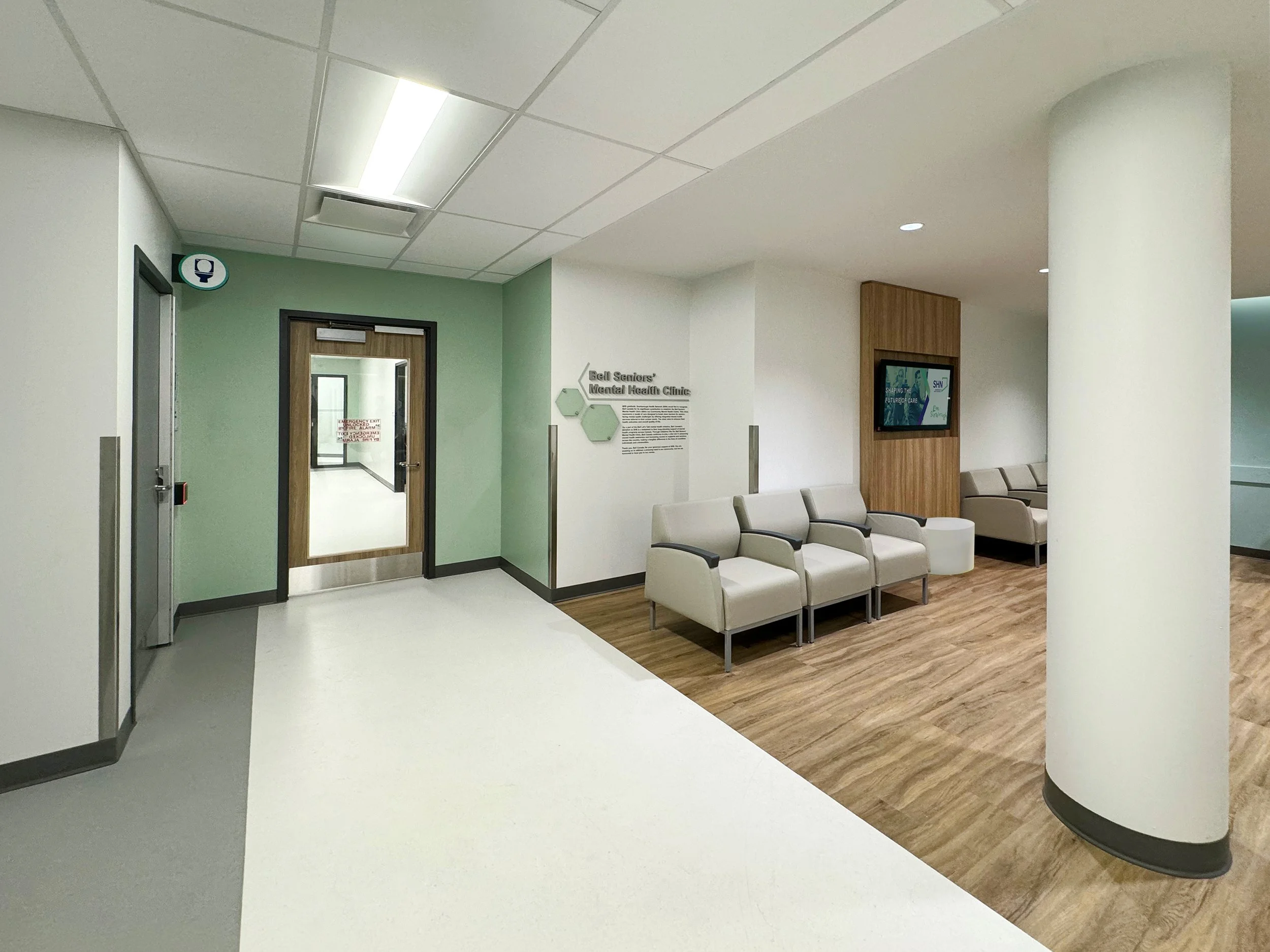 Waiting room area in a medical facility with white and beige chairs, digital screens, and a door leading to multiple rooms.