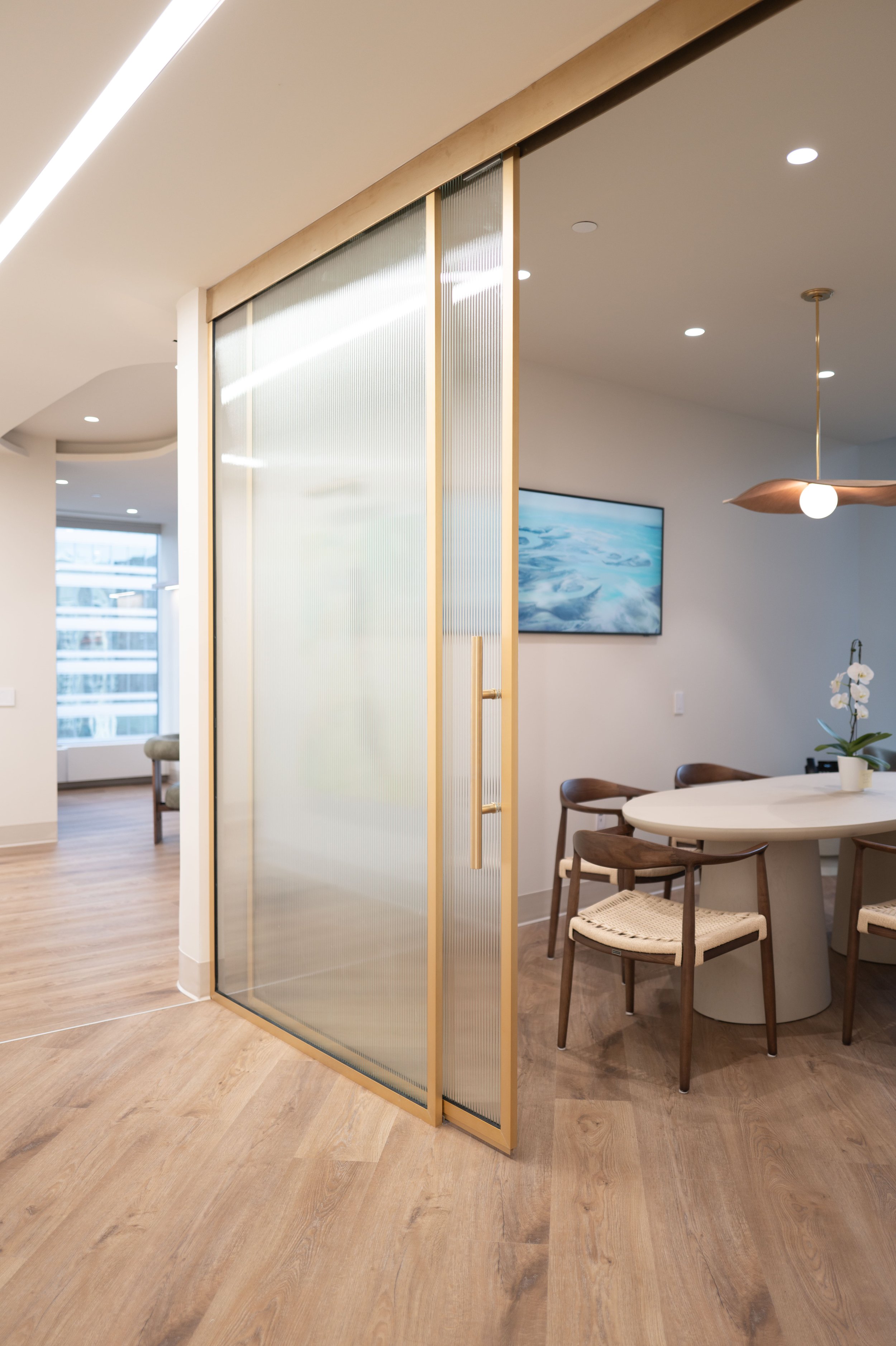 Modern dining area with round table, four chairs, a hanging pendant light, and a wall-mounted TV. A frosted glass sliding door separates it from the living space, which has large windows and a wooden floor.