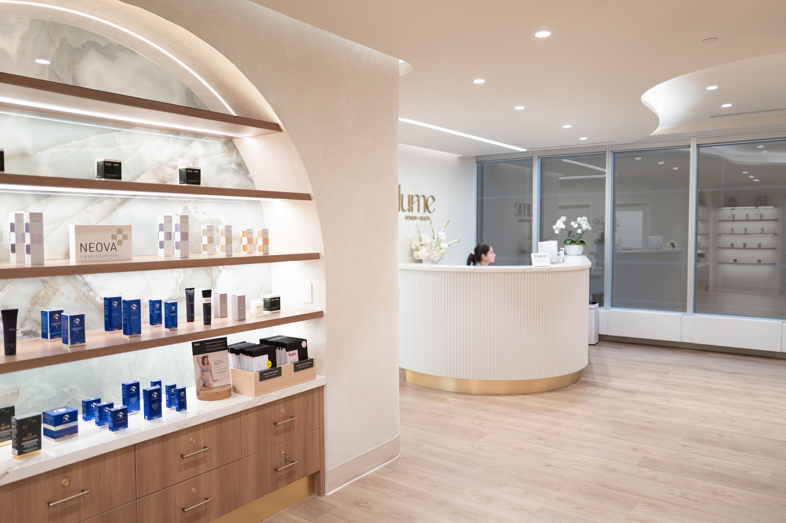 Modern spa reception area with a curved white desk and a woman working, shelves with skincare products, and large glass windows.