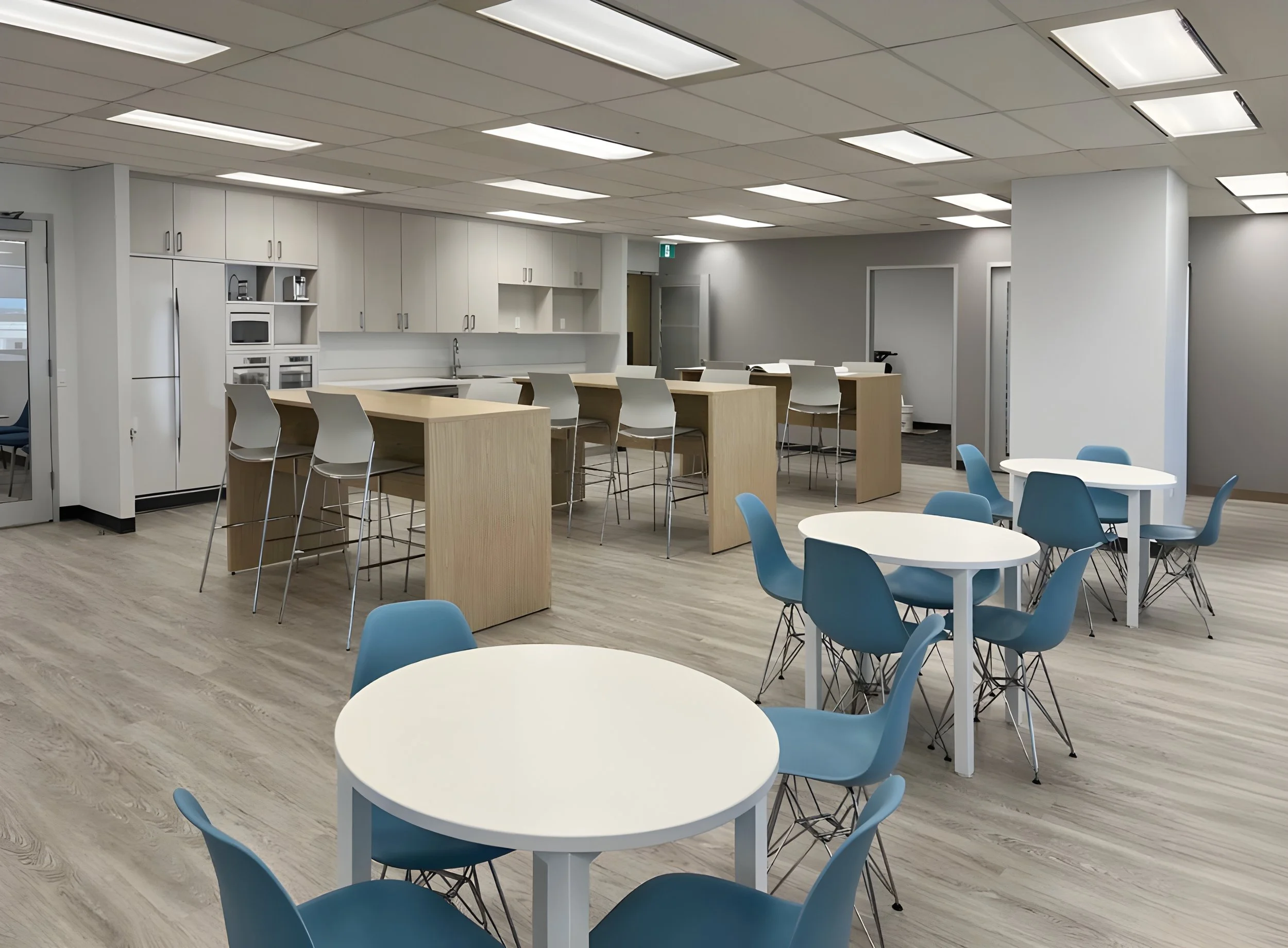 An office break room with white cabinets, a microwave, and a refrigerator along one wall. There are chairs around white round tables and light wood high-top tables,  and light wooden flooring. Toronto, Dallas, Texas, Miami, and Park city.