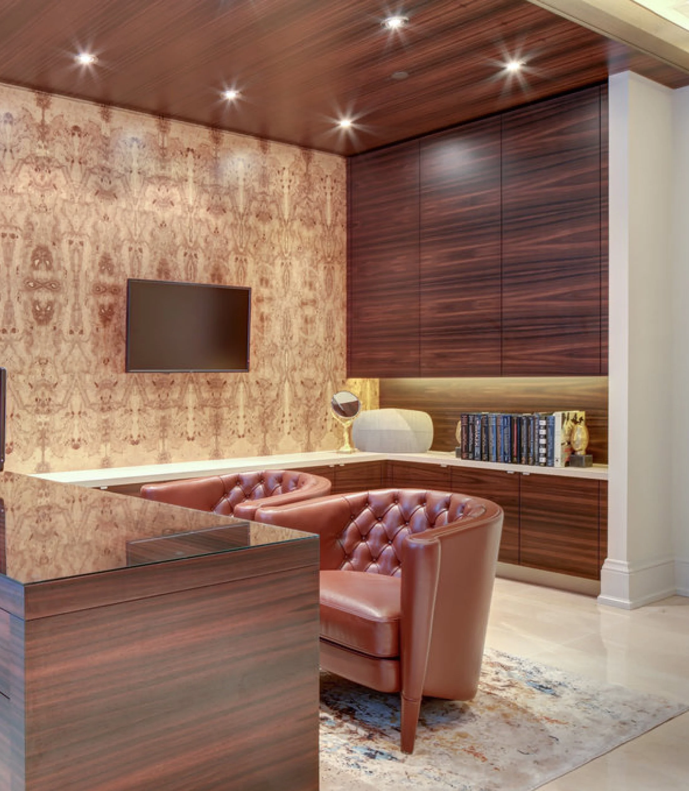 Modern living room interior with wooden ceiling, built-in wooden wall cabinets, a flat-screen TV, a chest of drawers, a wall-mounted mirror, a bookshelf, a gold decorative piece, a table lamp, leather armchairs, a patterned rug, and hardwood flooring
