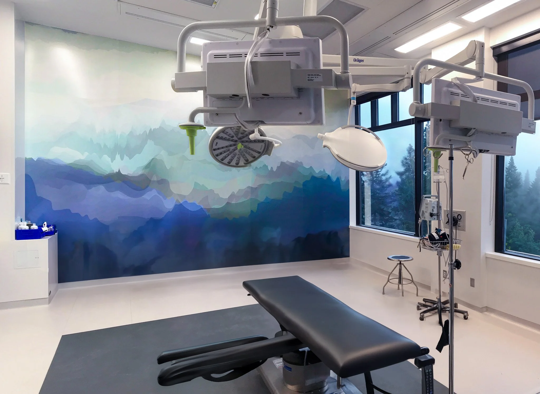 A hospital examination room features a medical chair, an adjustable overhead surgical light, and medical equipment. The room has a large window with a colorful mountain mural on the wall. Good inspiration for Toronto, Dallas and Park City.