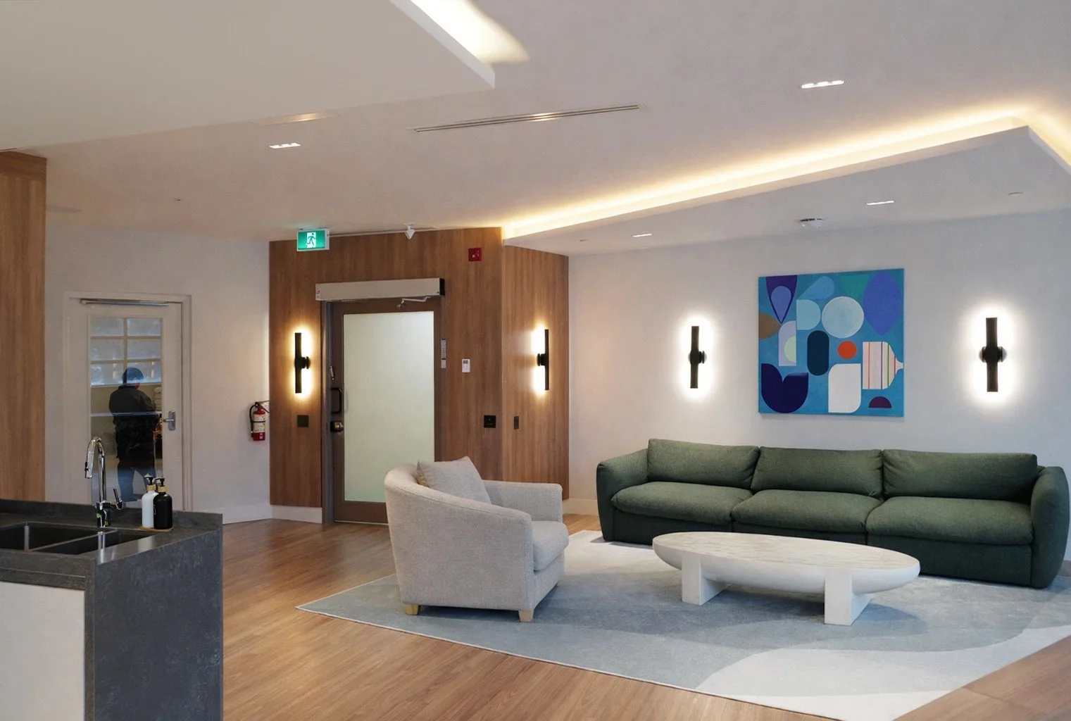 Modern lobby with a green sofa, beige armchair, abstract artwork on the wall, wood paneling, and a circular coffee table. Includes wall-mounted lights, fire extinguisher, exit sign, glass door with a person outside, and a kitchenette with a sink.