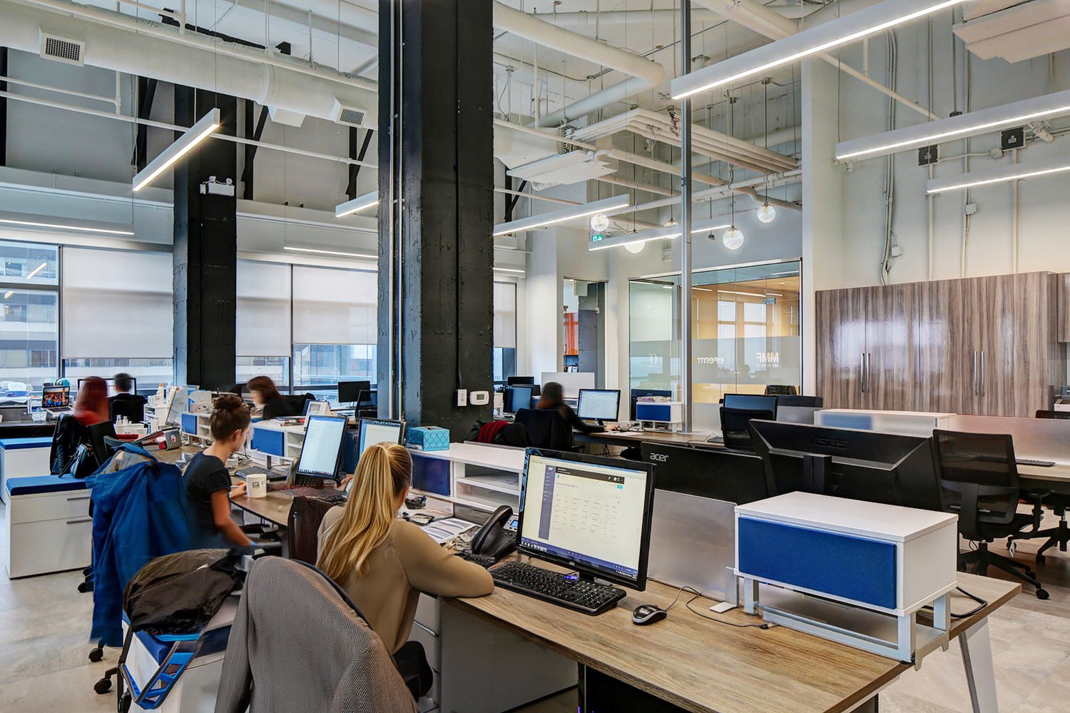 Open-plan modern office with multiple employees working at desks with computers, large windows with blinds, and contemporary lighting fixtures. Education Architecture. wellbeing and wayfinding. Inspiration for Toronto, Dallas, Park city, Texas, Miami