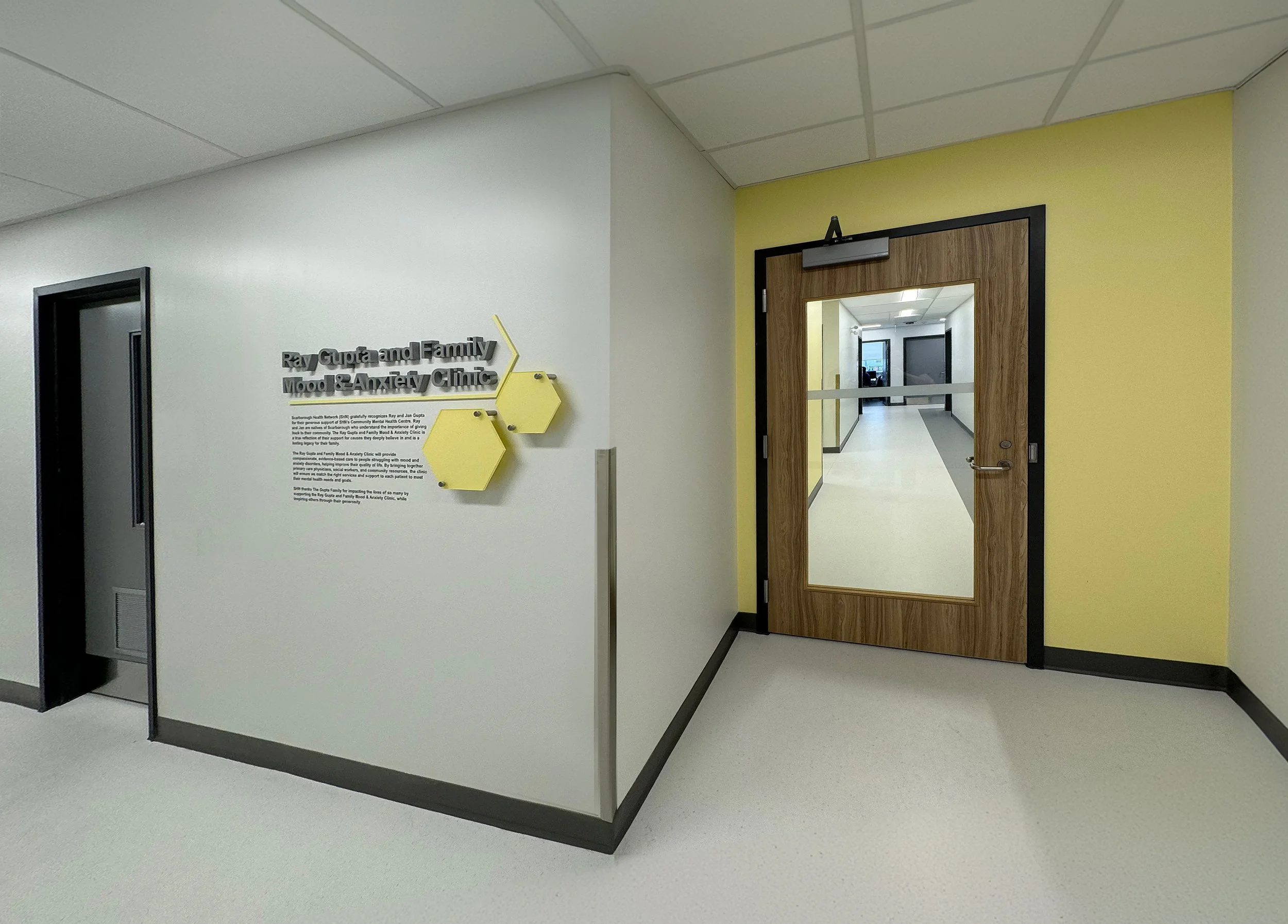 Hospital hallway with a yellow wall, wooden door with a mirror, and a sign indicating Ray Gupta and Family Mood & Anxiety Clinic