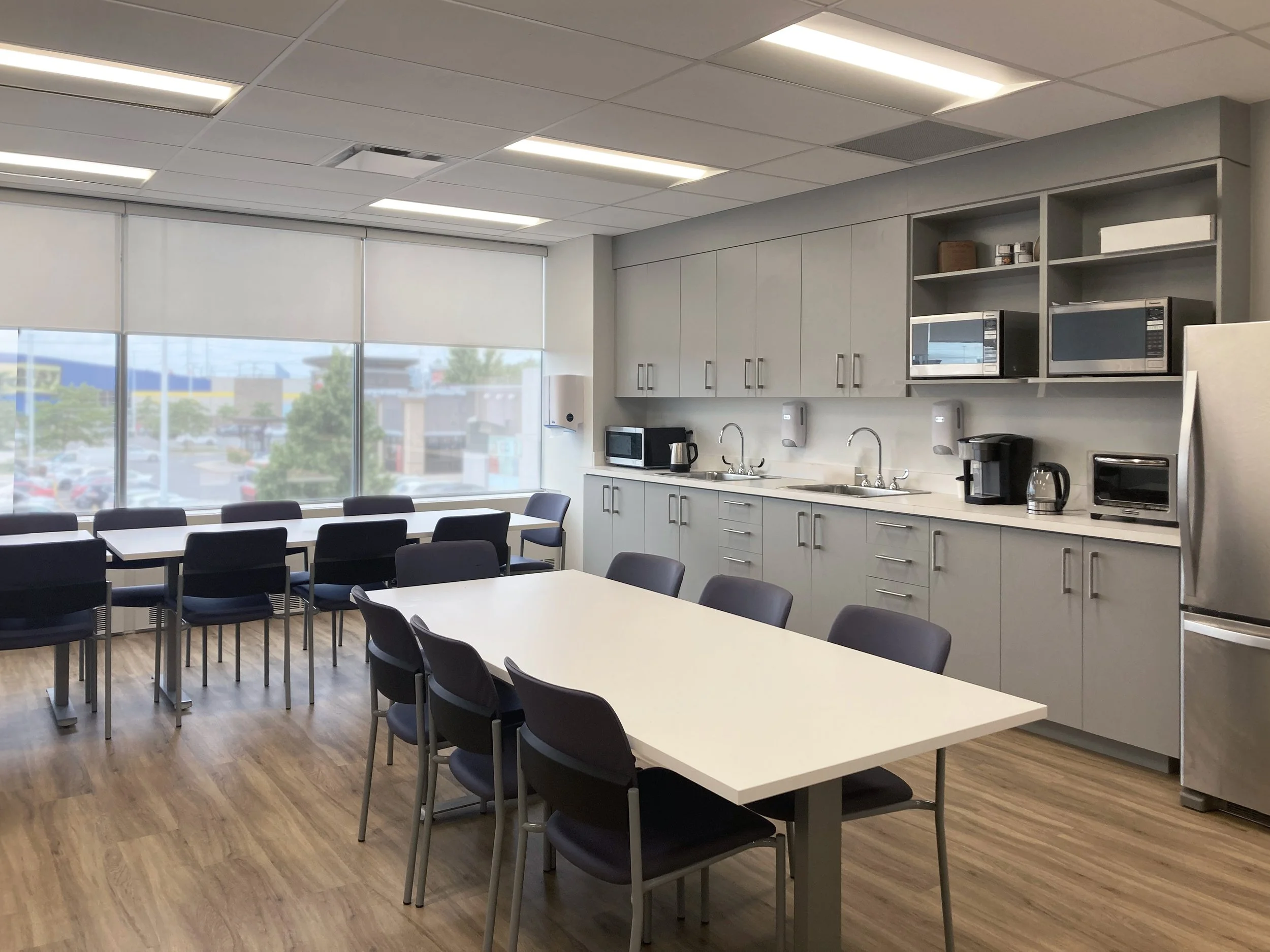 A modern office kitchen and break room with white cabinets, microwave ovens, and a countertop with appliances such as a coffee machine and kettle, along with tables and chairs for seating.