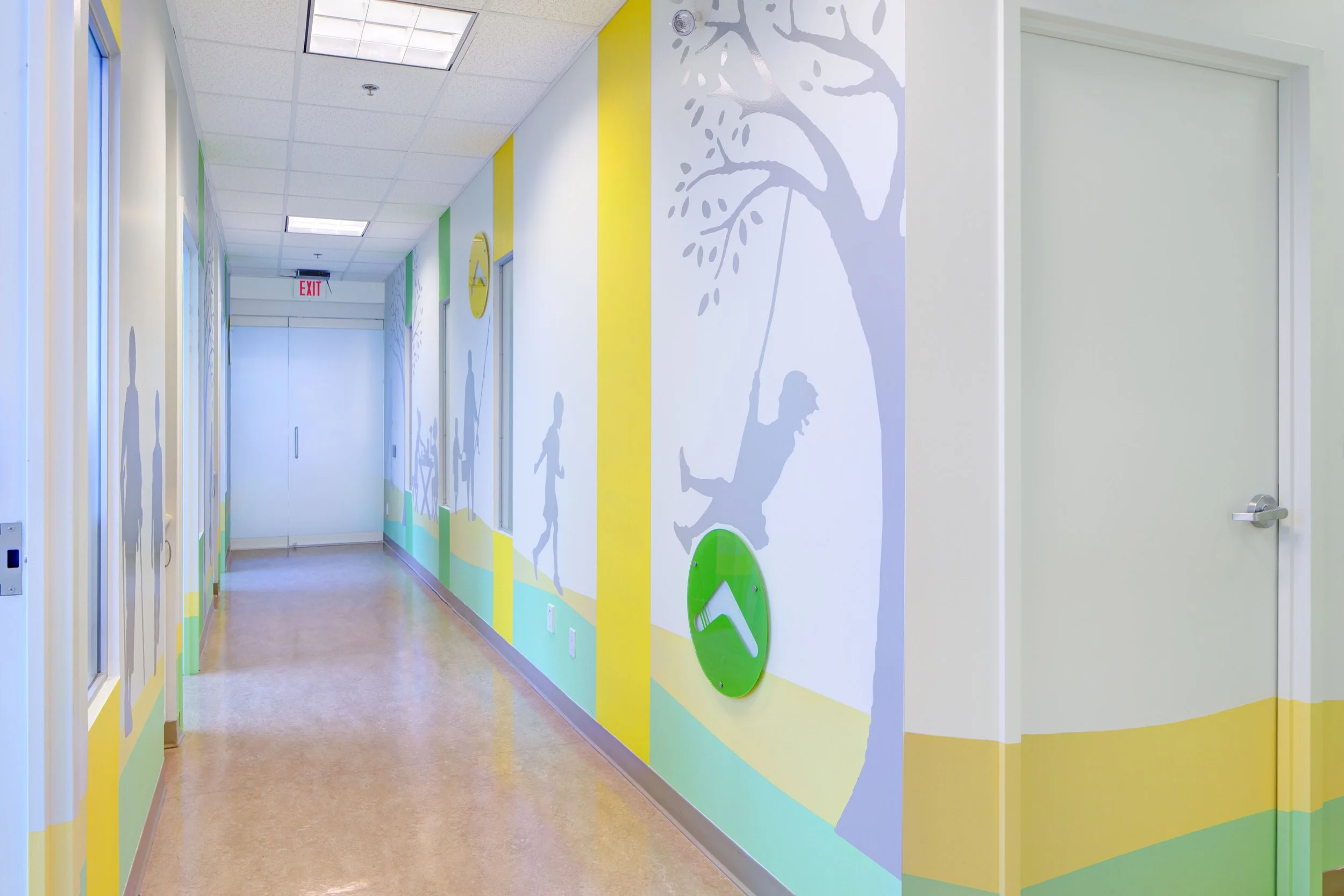 Colorful hospital hallway with murals of children playing and smiling. The walls are painted with bright yellow, green, and blue sections, and the ceiling has fluorescent lights. Healthcare. Inspiration for Toronto, Dallas, Texas, Miami and Park city