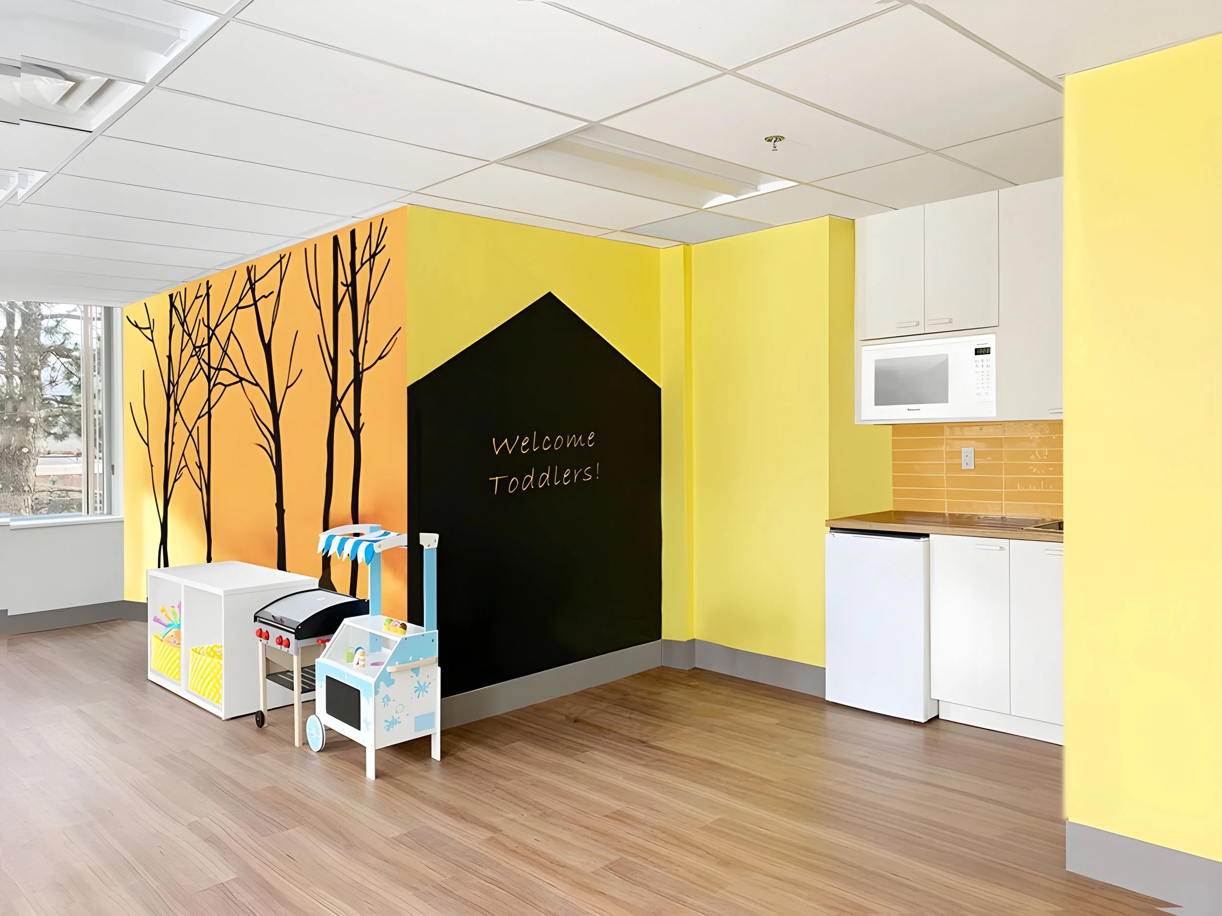 Colorful indoor space designed for toddlers, with a black accent wall featuring trees and welcome message, white toys, and yellow walls with white cabinets, microwave, and windows.