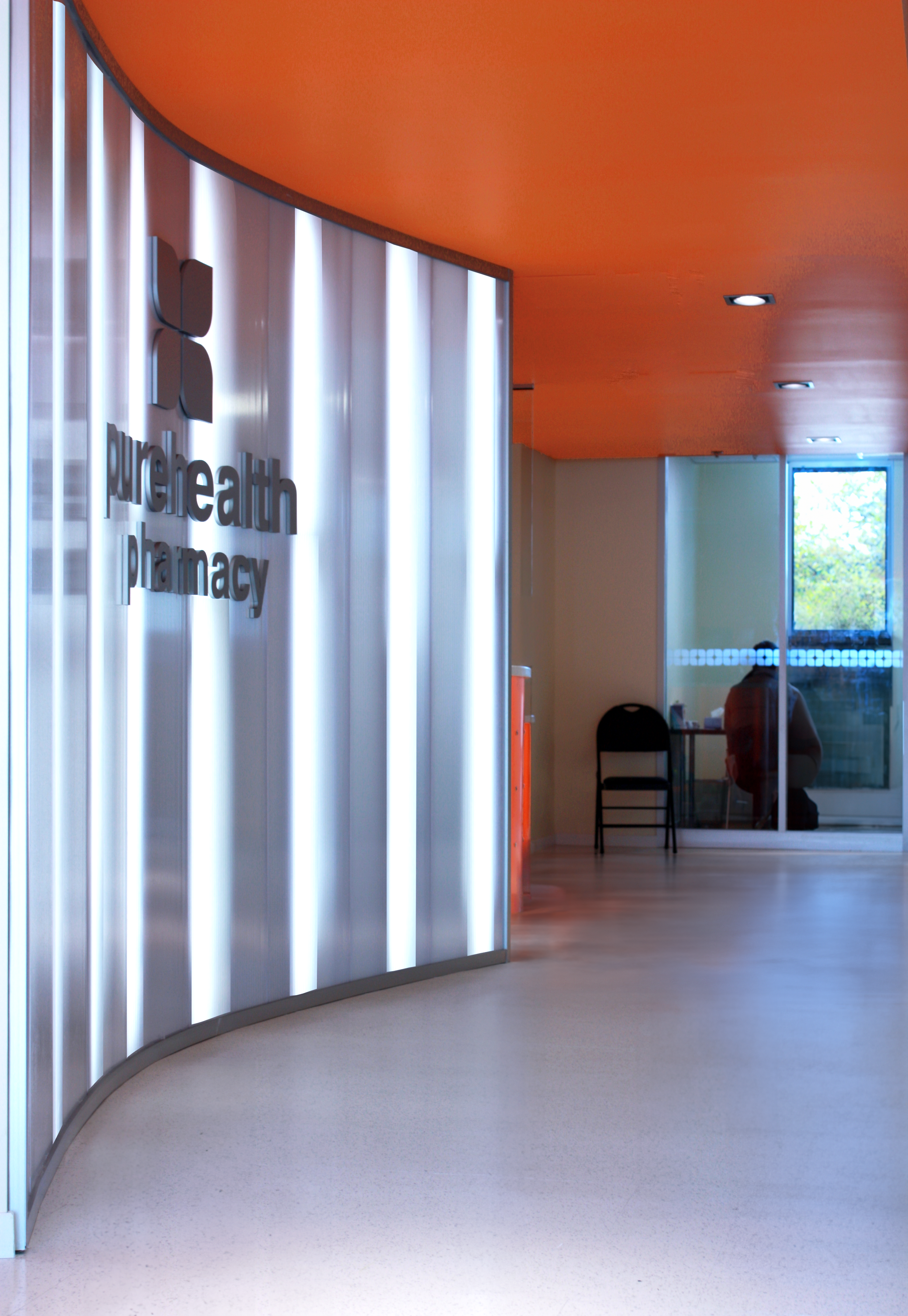 Interior view of a modern pharmacy with a white illuminated sign that reads 'purehealth pharmacy' on a curved wall. The ceiling is orange with built-in lighting. Healthcare, wayfinding, wellbeing. Toronto, Dallas, Texas, Miami and Park city.