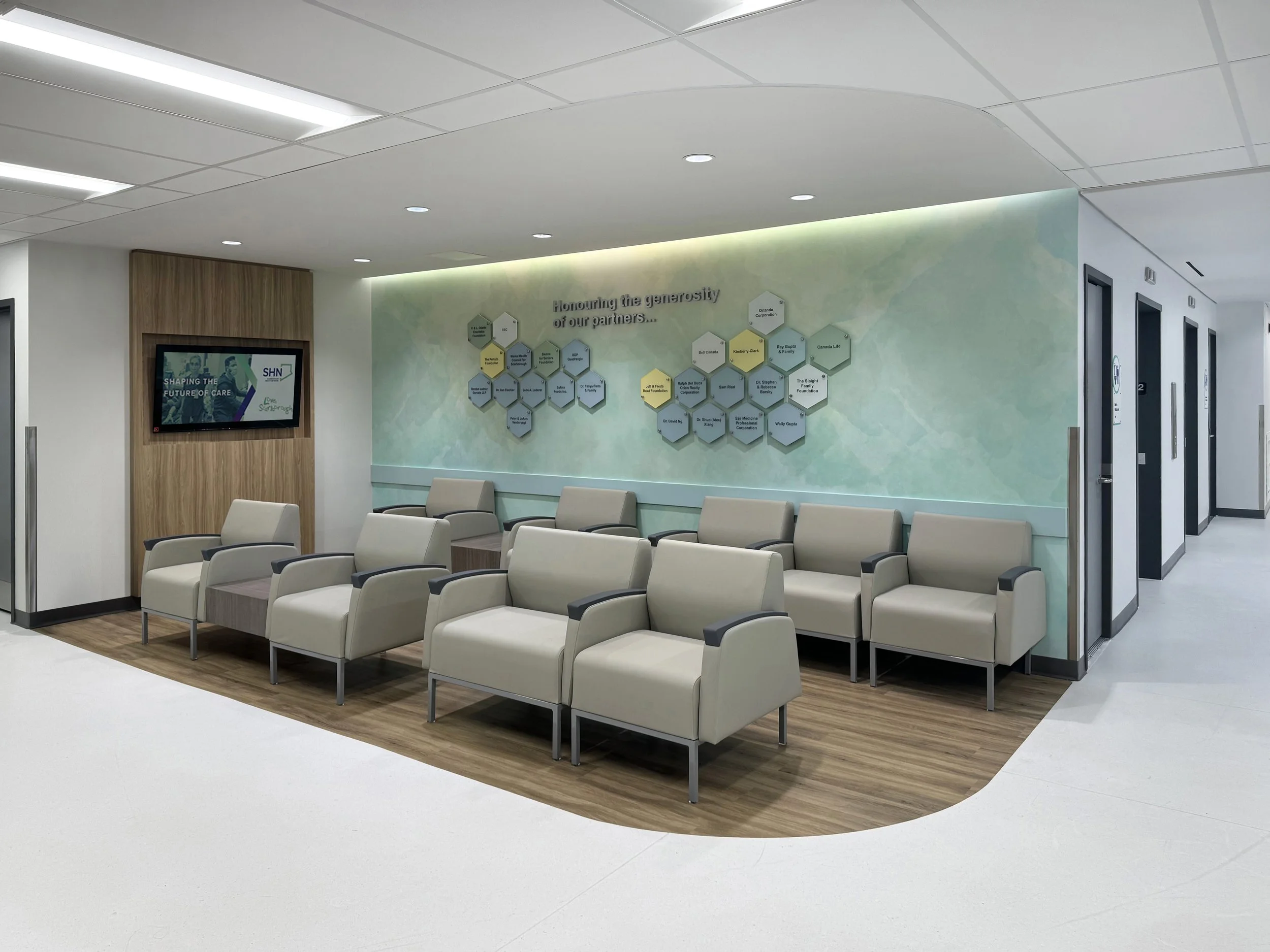 Hospital waiting area with beige chairs arranged in a semi-circle on a wooden floor, a wall with a greenish hue displaying a list of partners, and an electronic monitor showing a healthcare message.