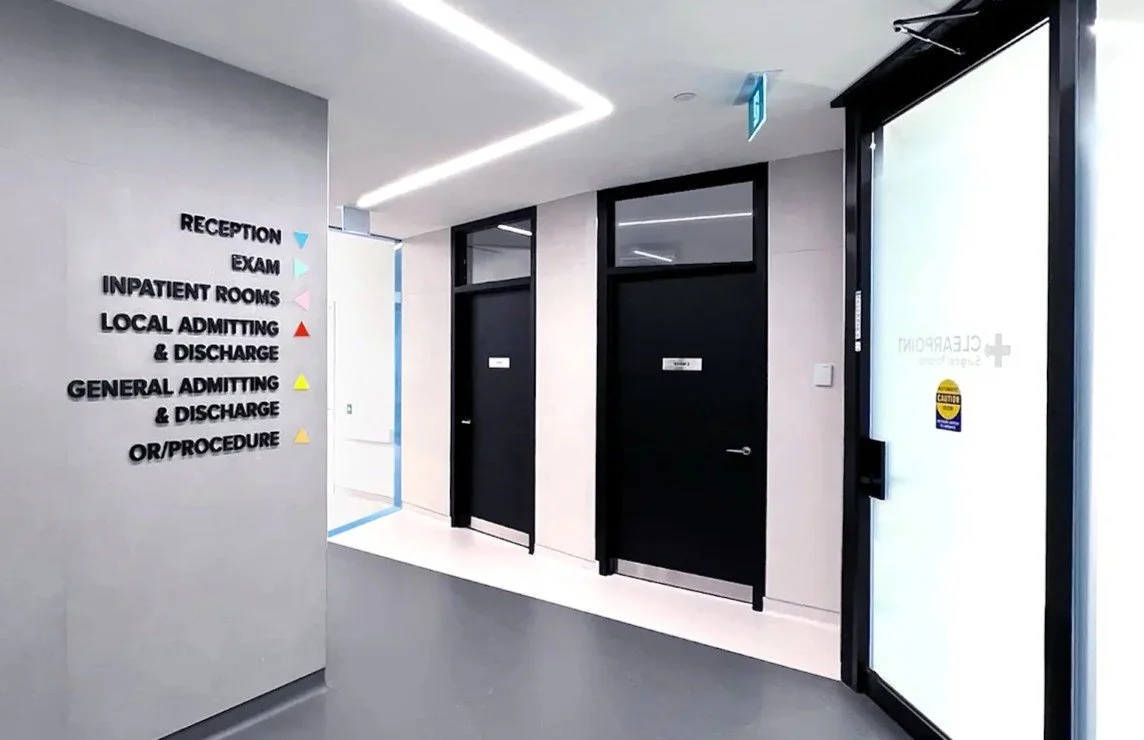 Hospital corridor with signs indicating directions to reception, exam, inpatient rooms, local admitting and discharge, general admitting and discharge, with white sliding glass door on the right. Good inspiration for Toronto, Dallas and Park City.