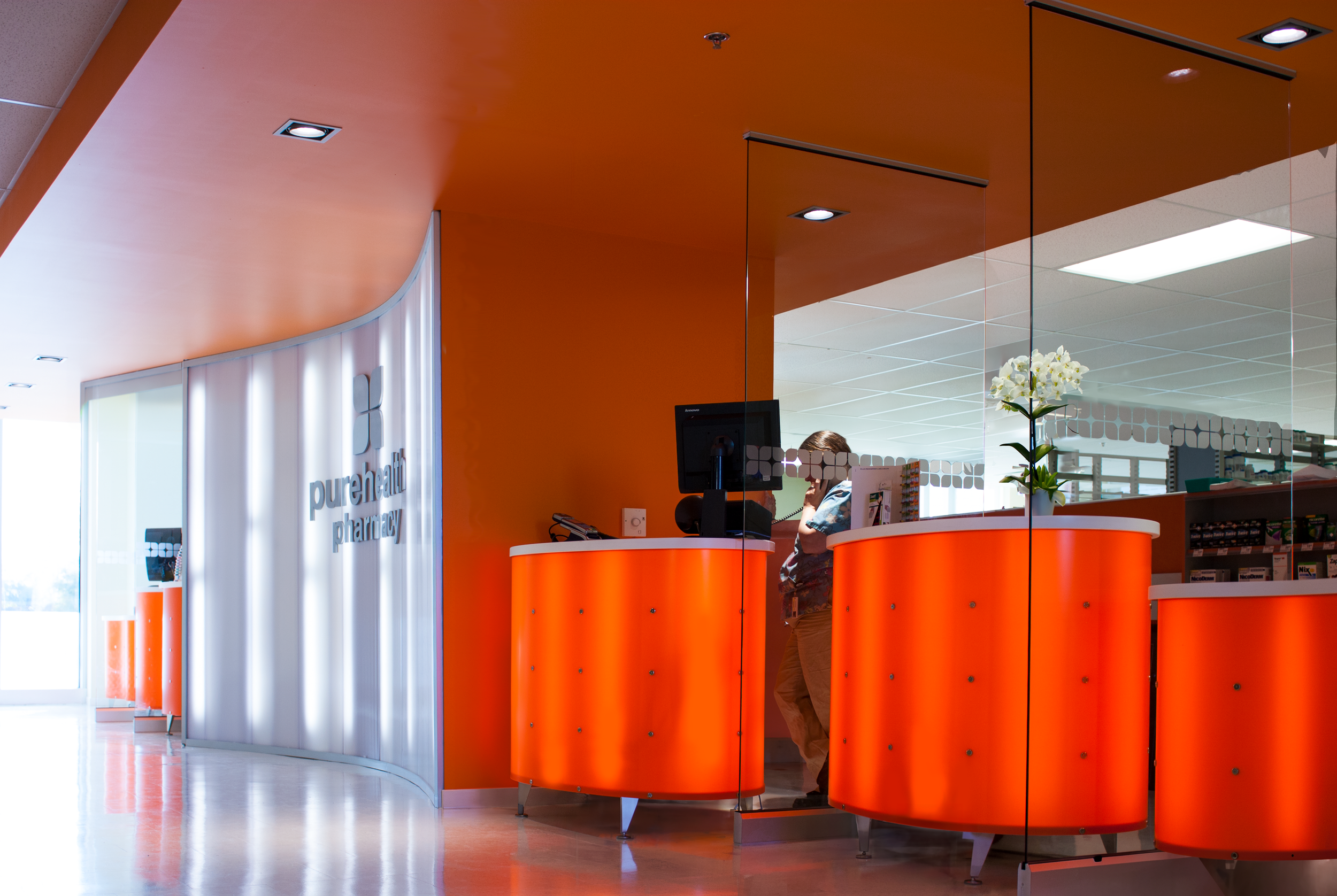Interior of a pharmacy with orange counters and a glass wall mirror. A person is behind the counter talking on the phone and another person is visible, partially obscured, behind the counter. healthcare. Toronto, Dallas, Texas, Miami, and Park city.