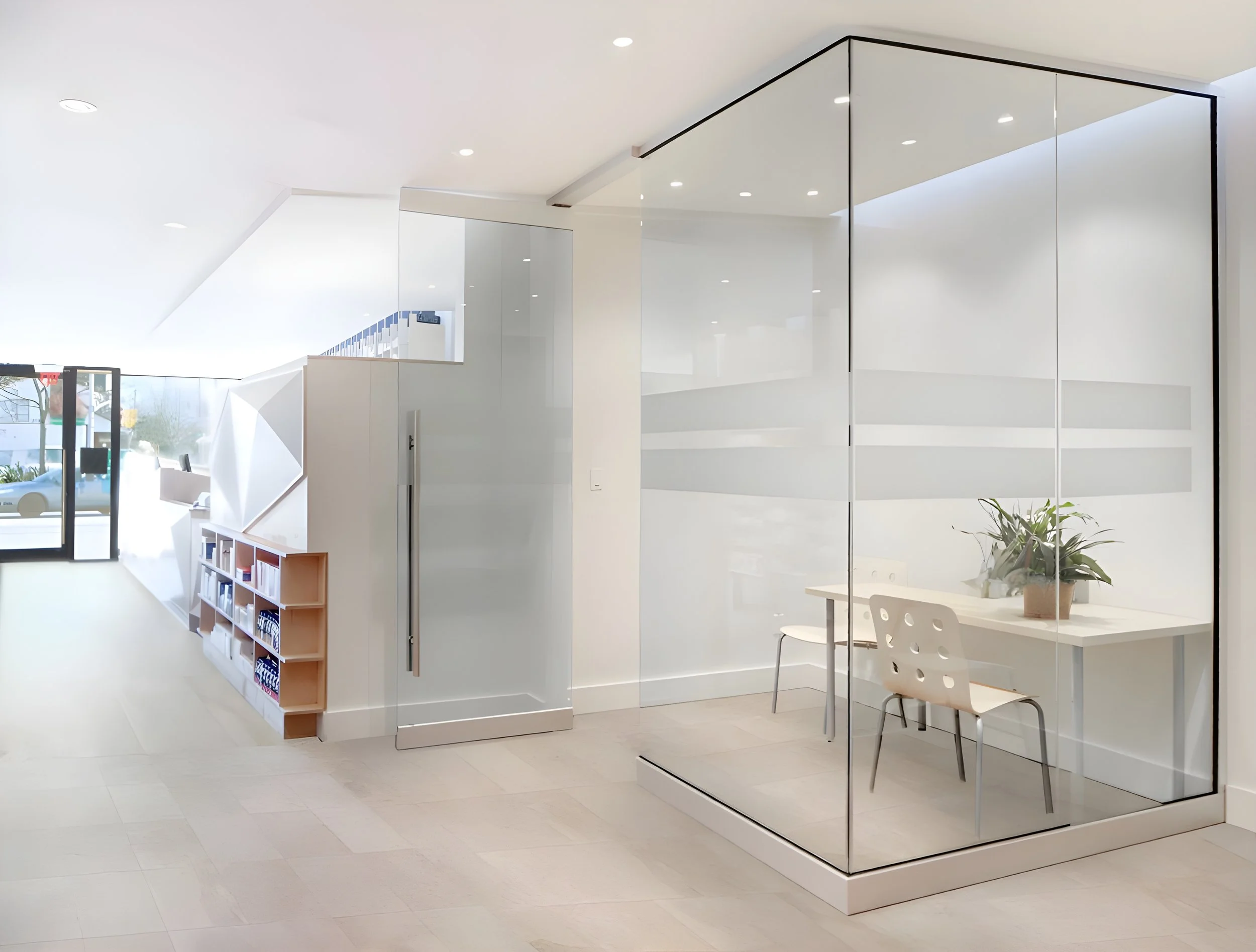 Modern interior of a bright, white office space with a glass wall, a small table with a chair, and a potted plant, with natural light coming through windows.
