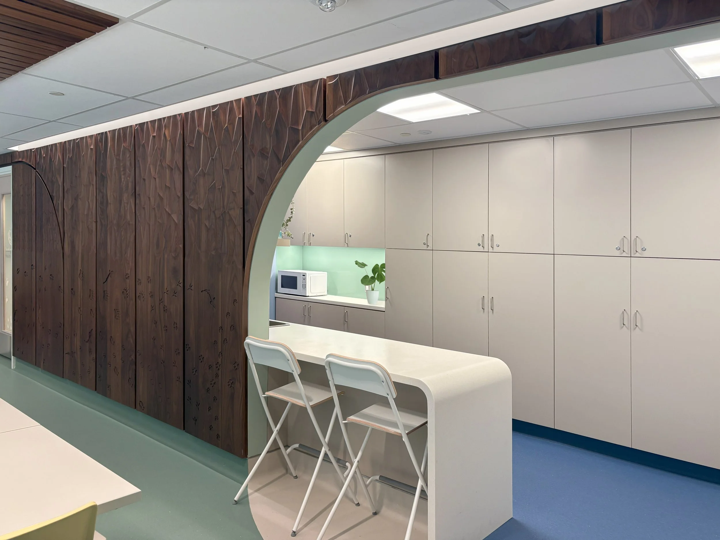 Interior view of a modern office or break room with a wooden decorative partition, white cabinets, a microwave, a potted plant, and chairs around a table.
