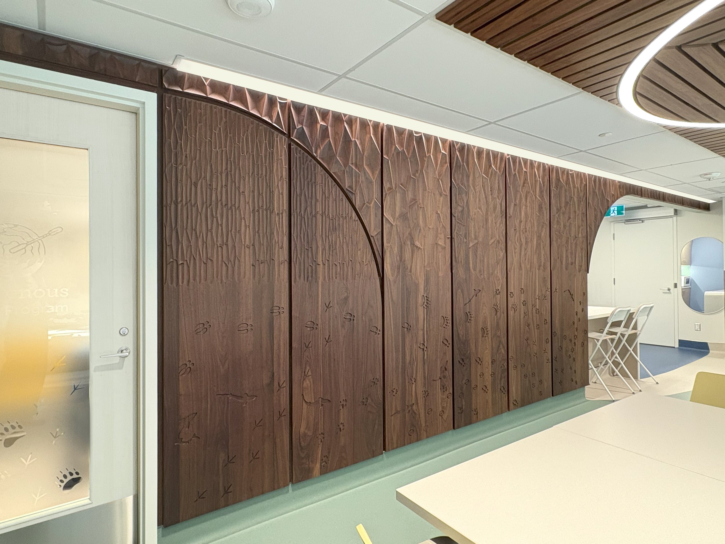 Wooden installation panels with engraved leaf and vine designs on an interior wall, against a ceiling with white tiles and a strip of LED lighting.