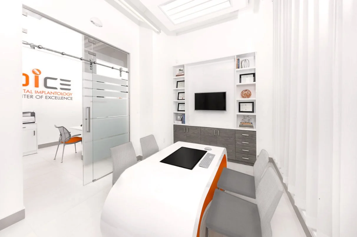Modern conference room with white walls, a white table, gray chairs, and a wall-mounted TV surrounded by white shelves and cabinets.