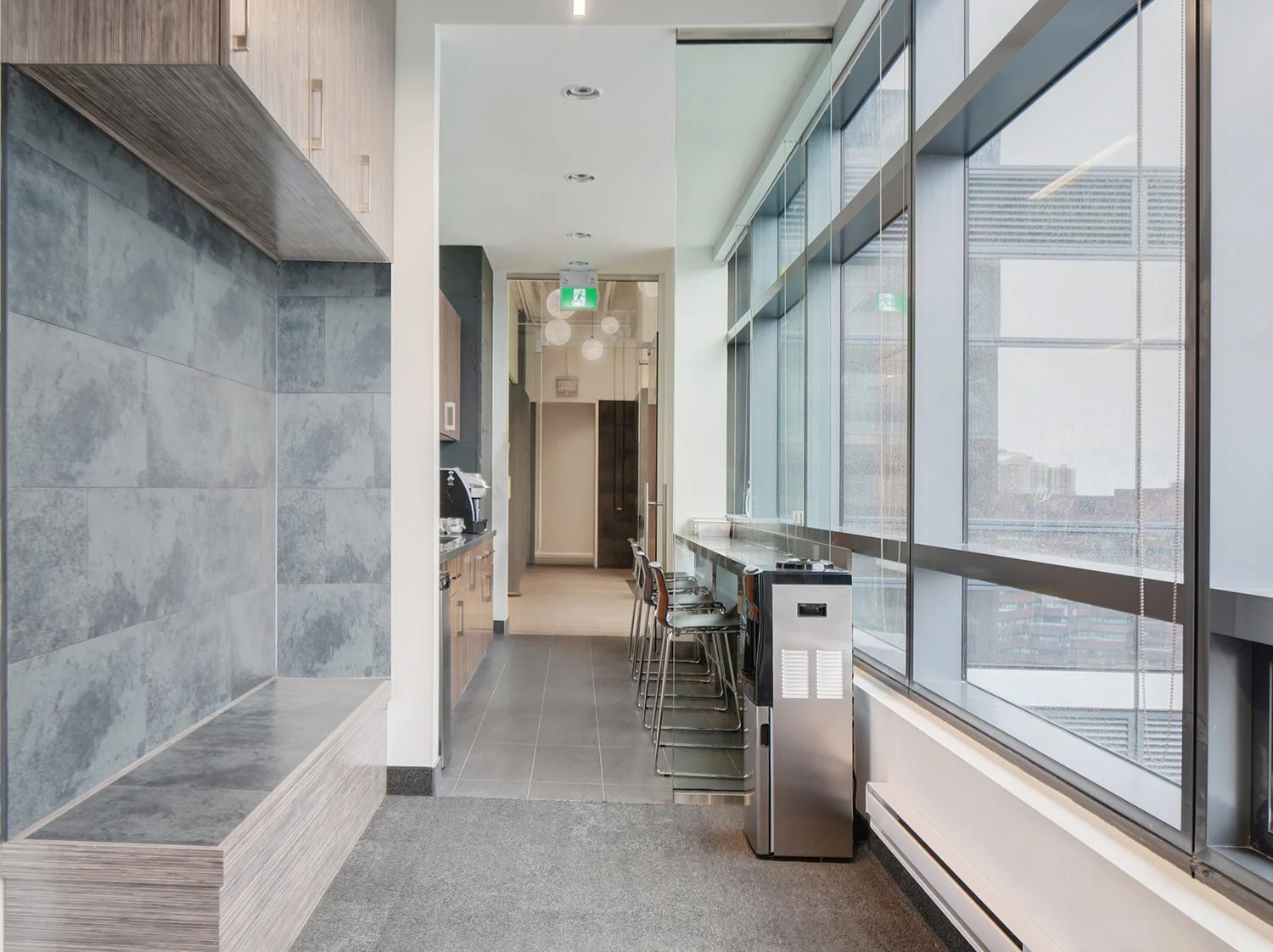 Indoor space with large windows, gray tiled surfaces, a bar-height counter with stools, and a coffee area with a microwave, suggesting a lounge or break room in a corporate or hotel setting. Education. Toronto, Dallas, Park city, Texas, Miami.