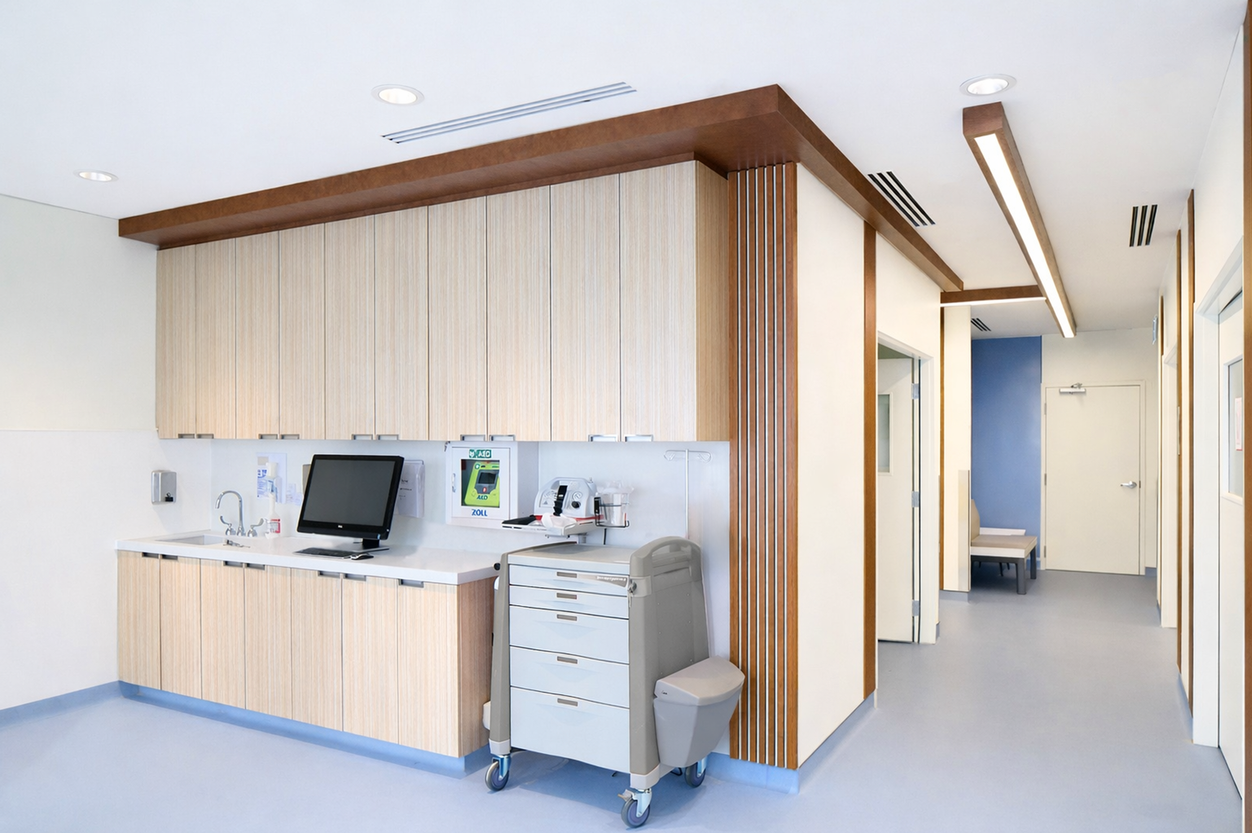 Hospital nurse station with wooden cabinetry, a computer, medical supplies, and a rolling cart, in a clinical setting with clean white walls and ceiling, and hallway with blue accent wall. Healthcare. Inspiration for Toronto, Dallas, Park city.