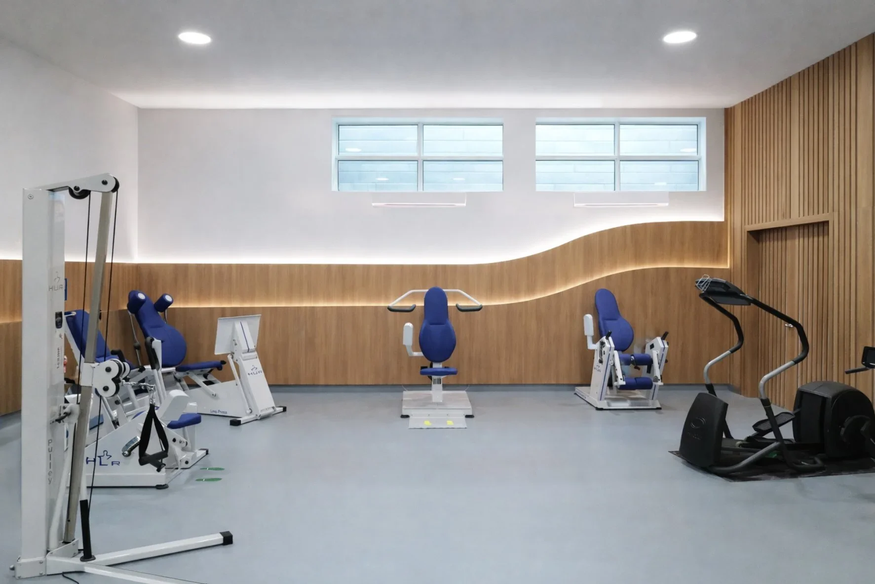 Modern indoor gym with gym equipment including leg press, rowing machine, and treadmill, featuring wooden wall accents and high windows.