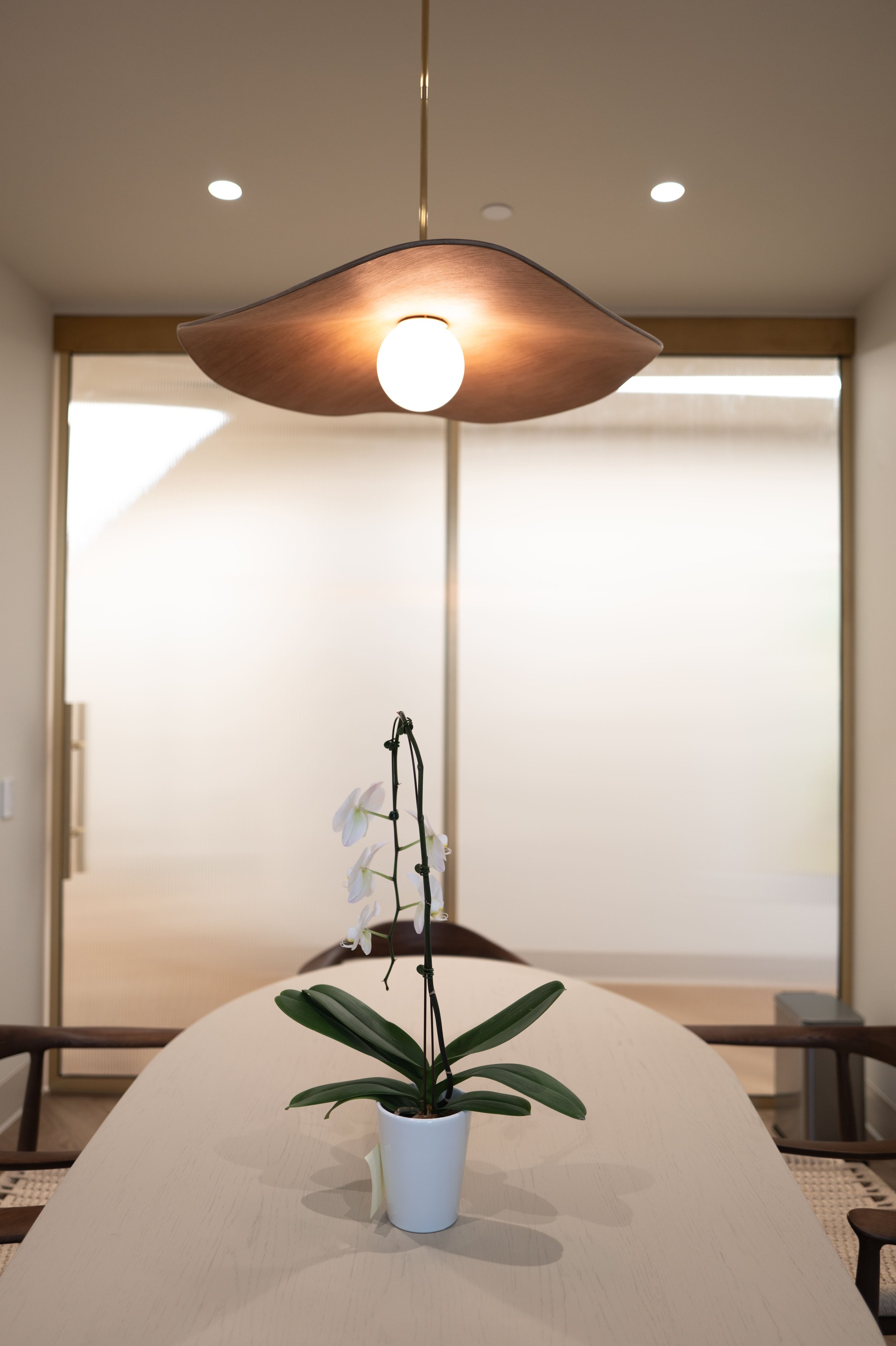 A dining room featuring a hanging ceiling light, a potted orchid on a table, and a background of large, frosted windows.