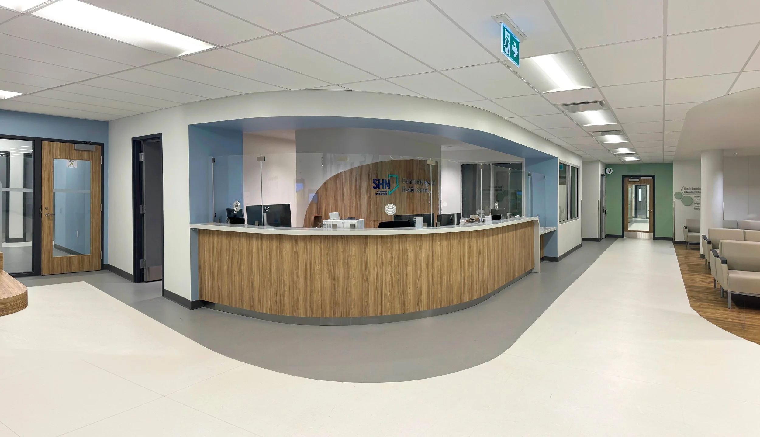 Hospital reception area with a curved wooden front desk, seating area with chairs, doors, signs, and a hallway.